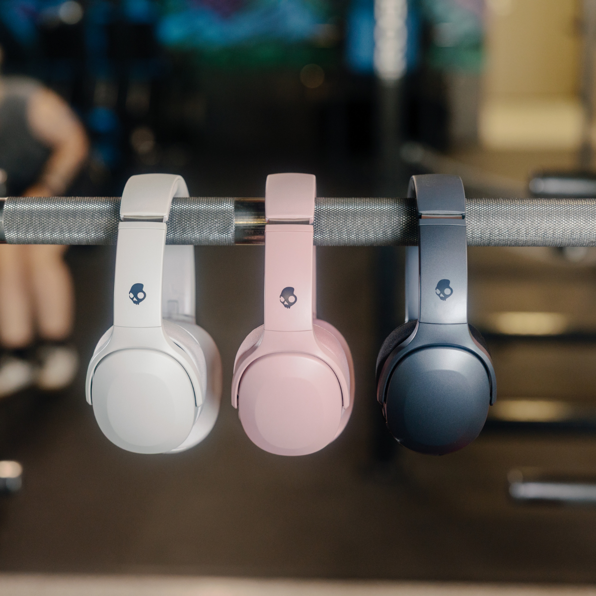 Trzy słuchawki wiszące na metalowym drążku. Białe, różowe i szare. Tło siłowni.