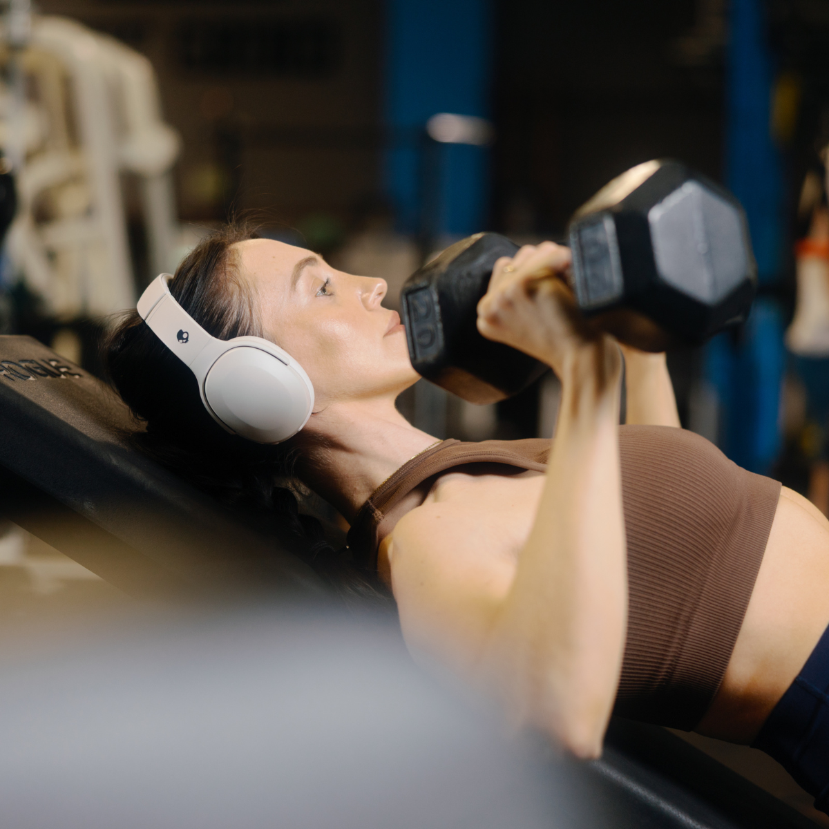 Kobieta na siłowni, podnosząca hantle w białych słuchawkach. Tło siłowni.