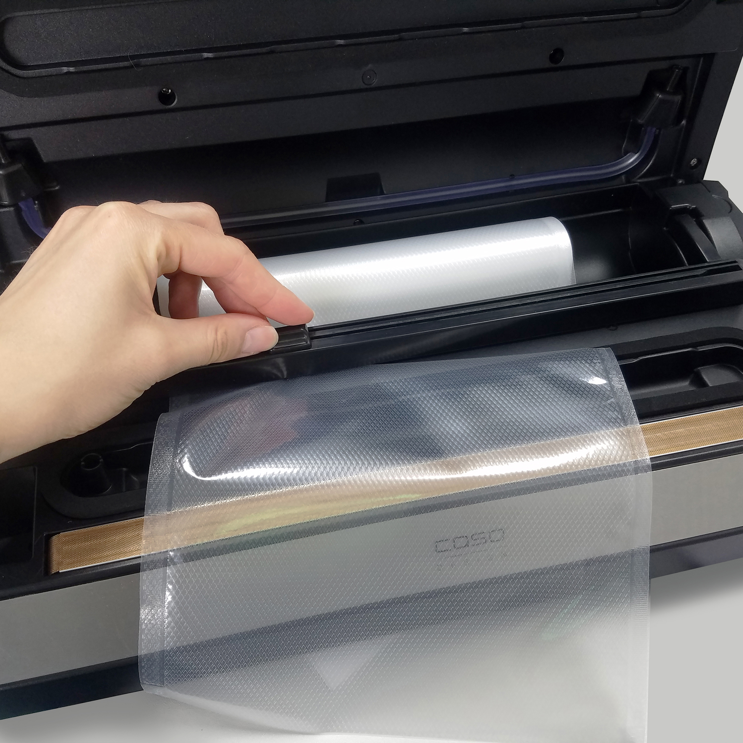 Eine Hand legt eine Plastikrolle in einen Caso-Vakuumierer auf einer weißen Oberfläche.