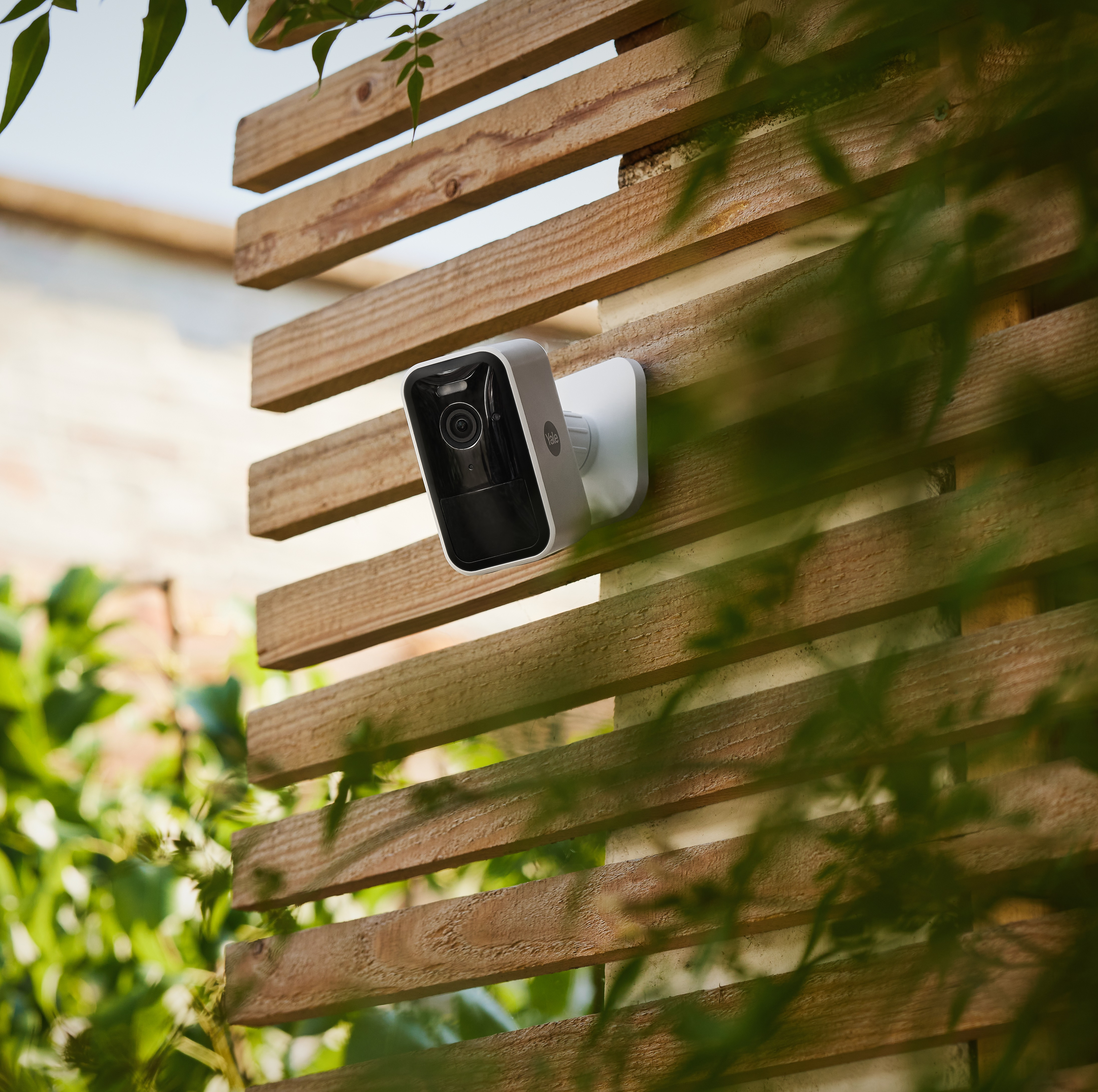 Biała kamera bezpieczeństwa zamontowana na drewnianym płocie, częściowo zasłonięta przez zielone liście.