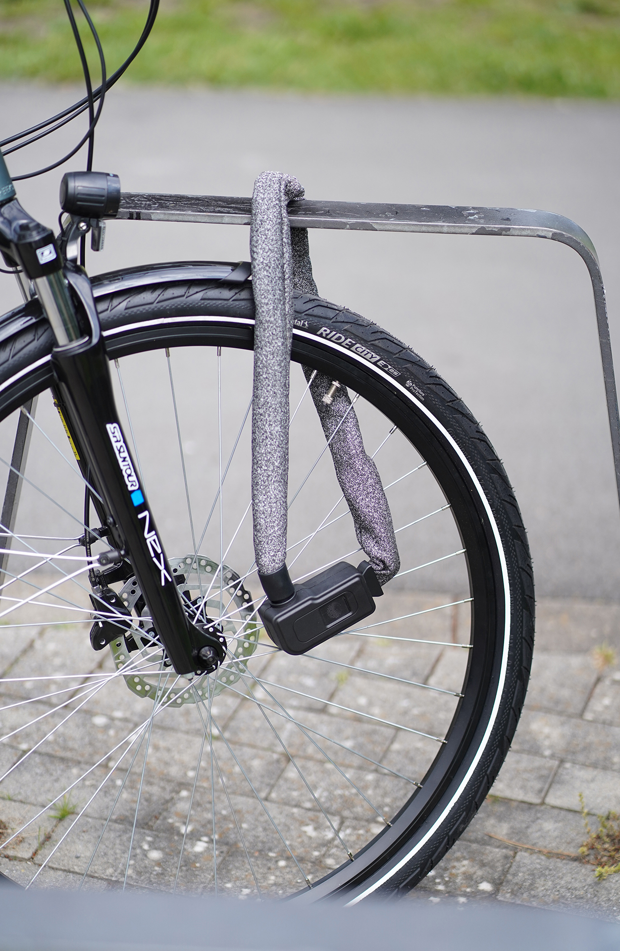 Ein Fahrrad mit Kettenschloss, das das Hinterrad an einem Gestell sichert. Das Fahrrad steht auf einer gepflasterten Fläche.