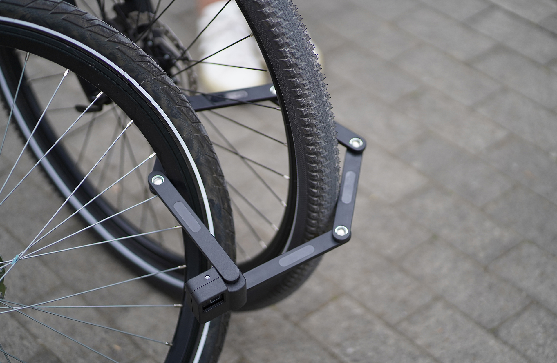 Schwarzes Fahrradschloss sichert zwei Fahrradräder. Grauer Pflasterhintergrund.