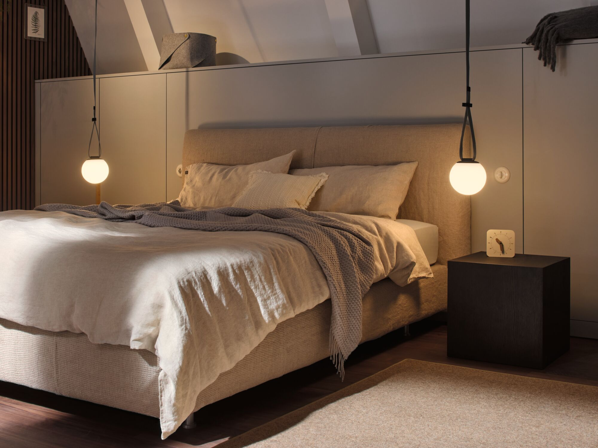 Schlafzimmer mit Bett, Nachttisch und zwei Hängelampen. Neutrales Farbschema.