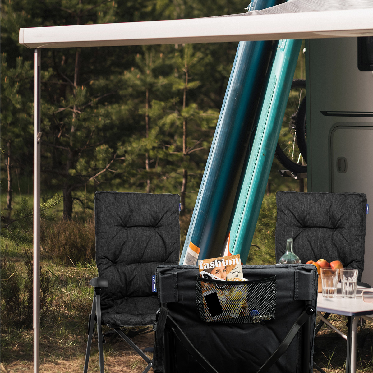 Camping scene with two chairs, a table, and a camper in the background. Trees are visible.