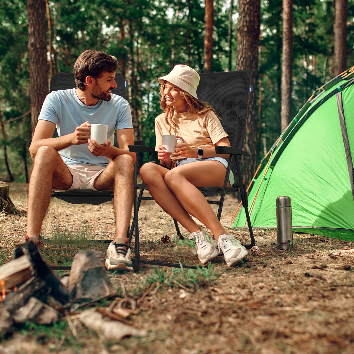 Een stel zit op stoelen, met koffie. Een tent en kampvuur op de achtergrond, in een bos.