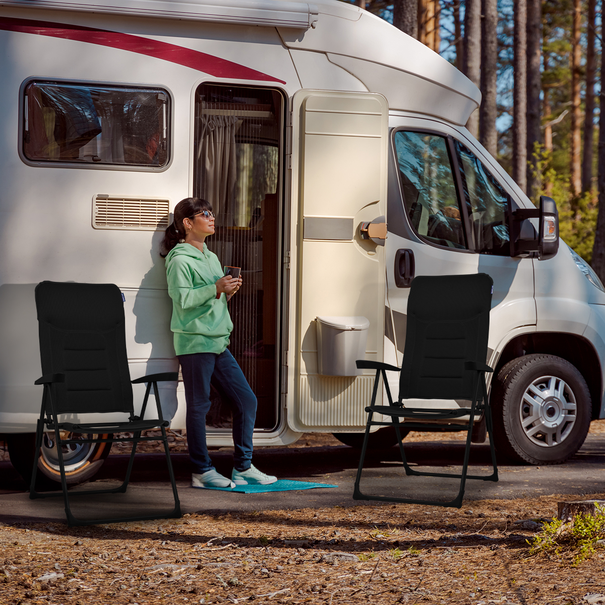 Een vrouw staat bij een camper met twee zwarte stoelen ervoor. Bomen zijn zichtbaar op de achtergrond.