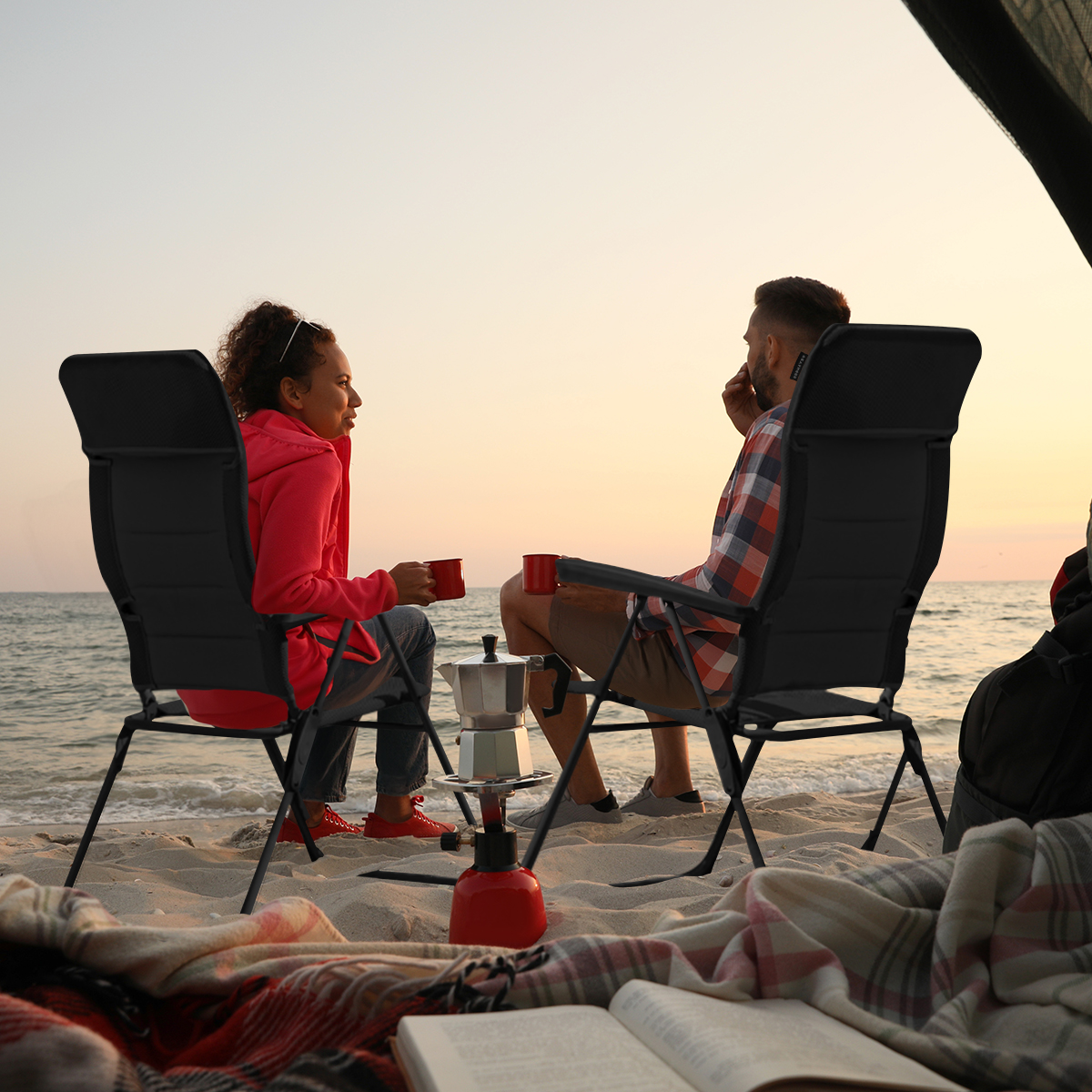 Een stel zit op stoelen op een strand, met een deken en koffiepot. De zee en zonsondergang zijn zichtbaar.