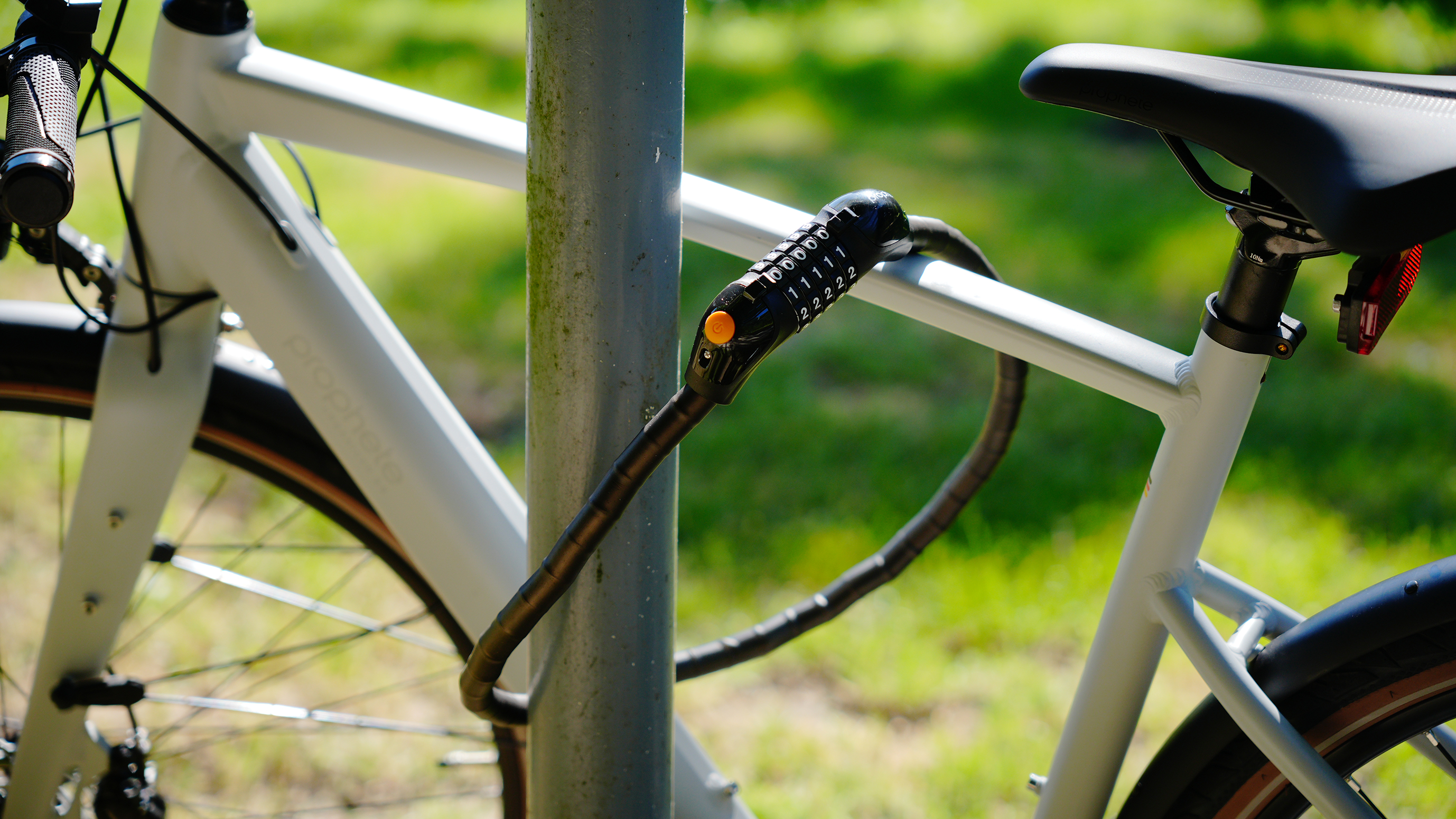 Ein weißes Fahrrad ist mit einem schwarzen Zahlenschloss an einem Metallpfosten befestigt. Grünes Gras im Hintergrund.