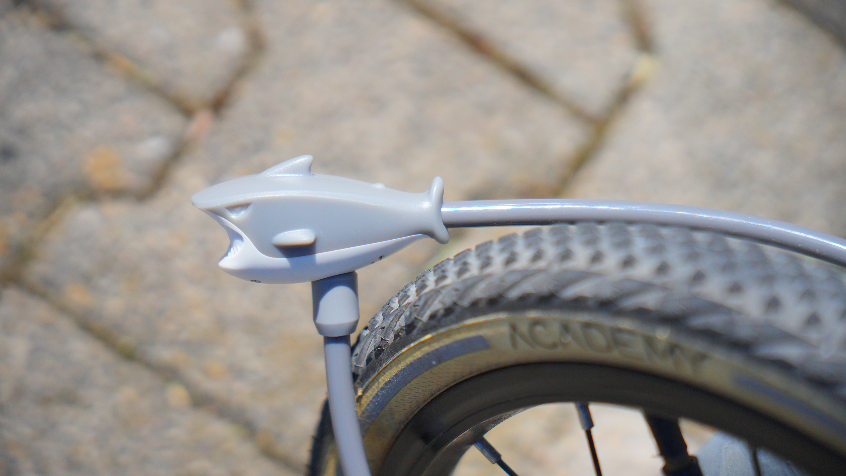 Ein Fahrrad-Schutzblech, verziert mit einem grauen Cartoon-Hai. Der Reifen ist sichtbar, das Pflaster grau.