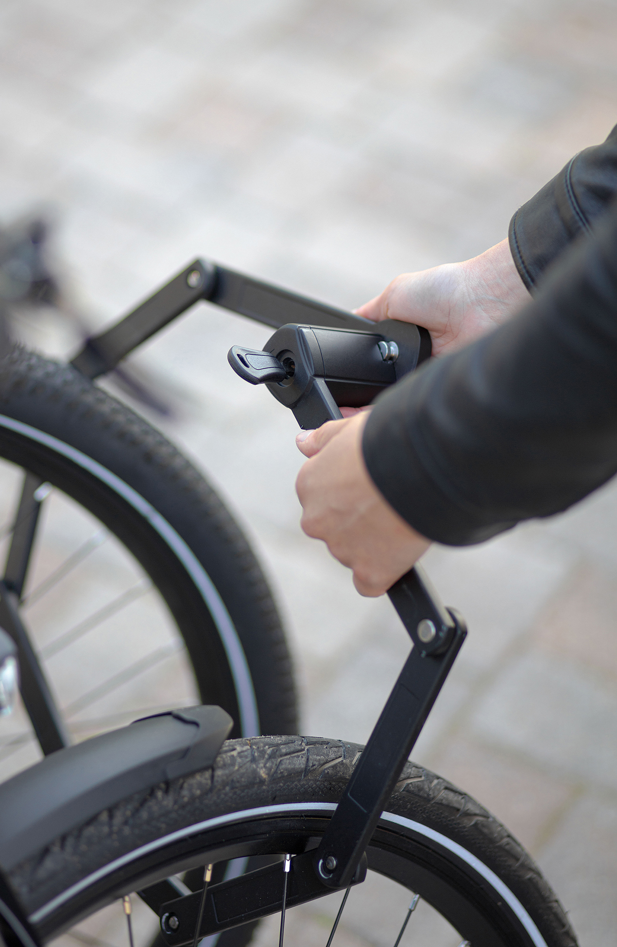 Eine Person hält ein schwarzes Fahrradschloss mit einem Schlüssel. Das Fahrrad steht auf einer gepflasterten Oberfläche, mit schwarzer Lederjacke.