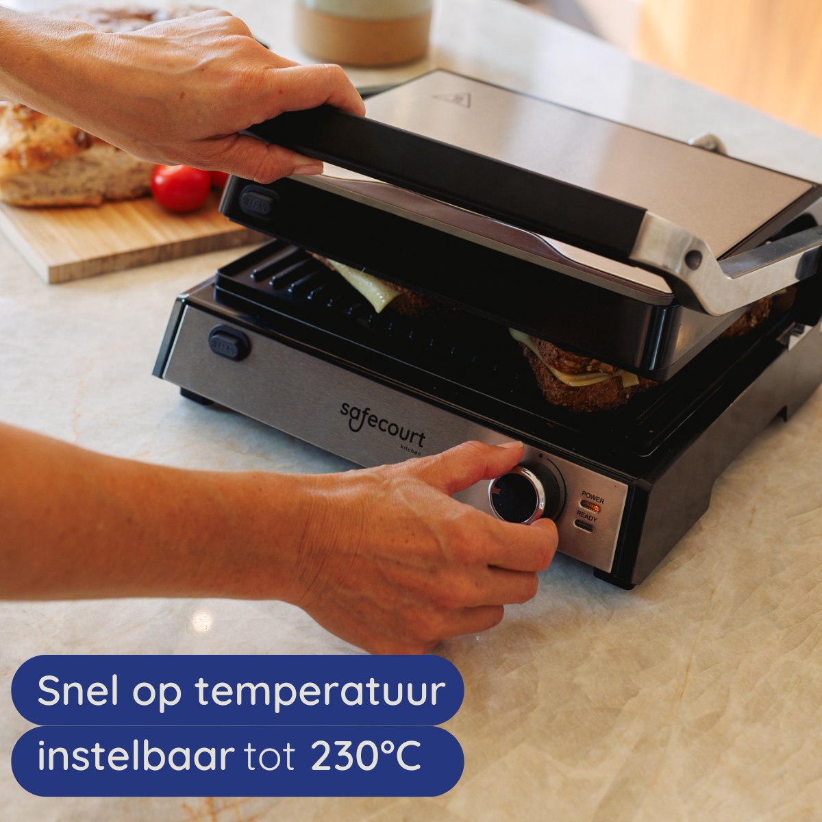 A person adjusts a grill's temperature dial. A sandwich is cooking on the grill. Bread and tomatoes are nearby.