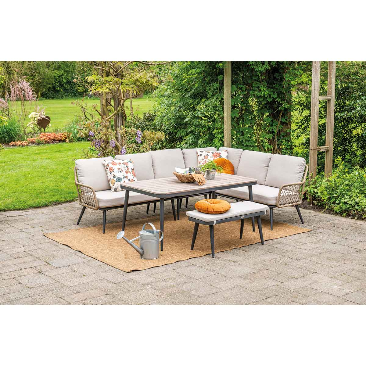 Canapé d'angle avec coussins gris, table en bois et banc sur un patio en pierre. Arbres en arrière-plan.