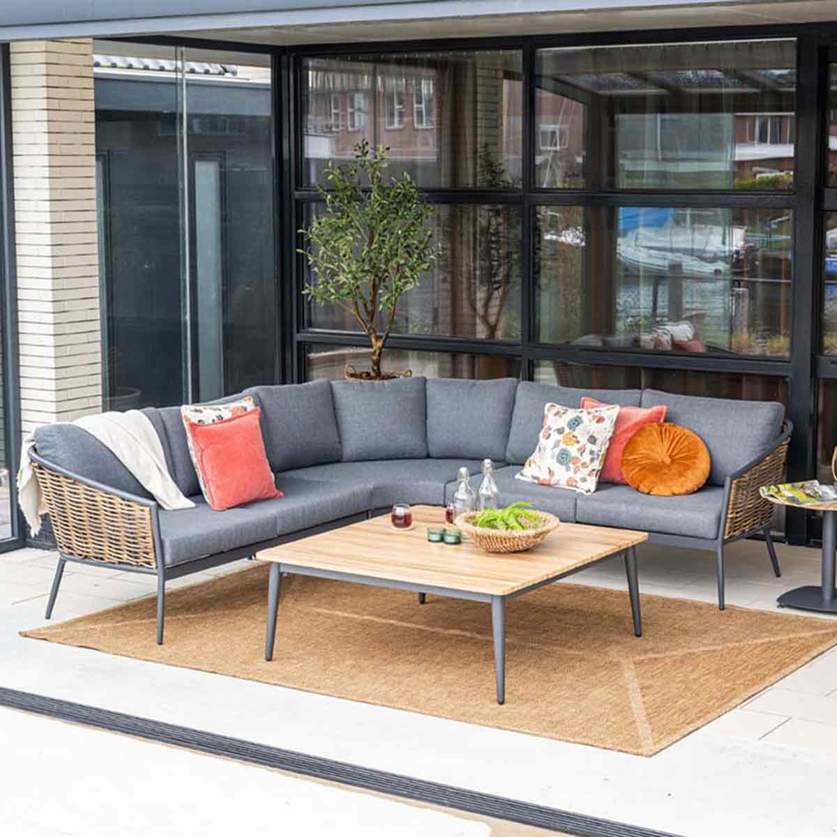 Outdoor sectional sofa with cushions, pillows, and a wooden table on a woven rug. Greenery and windows are in the background.