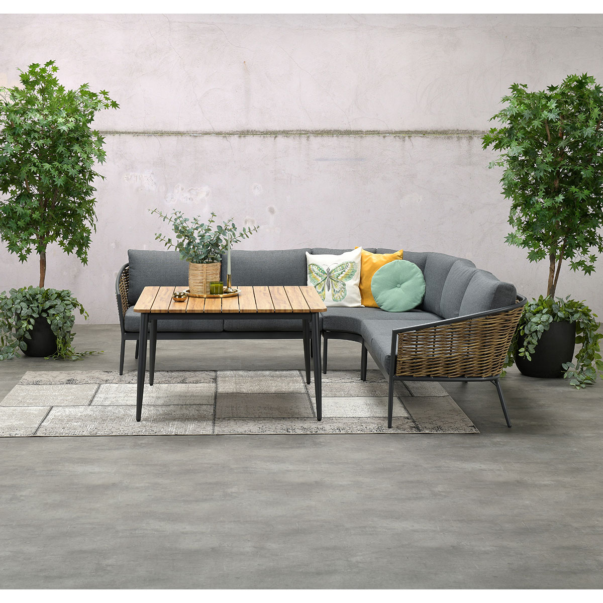 Canapé d'angle extérieur avec une table en bois, des plantes et un tapis sur un sol en béton. Des plantes vertes encadrent la scène.