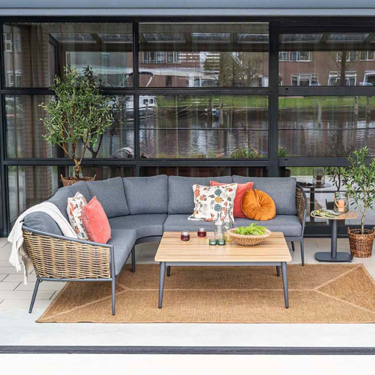 Outdoor seating area with gray sectional sofa, wooden table, and rug. Pillows, plants, and a view of water and buildings are visible.