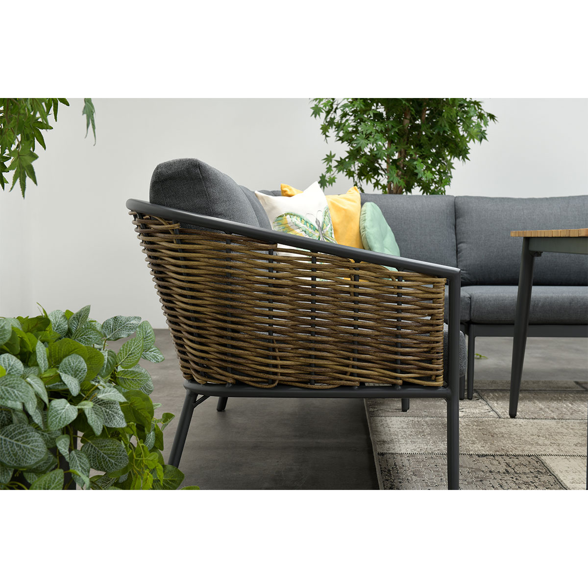 Gros plan sur un canapé d'angle extérieur avec un côté en osier, des coussins gris et une plante au premier plan.