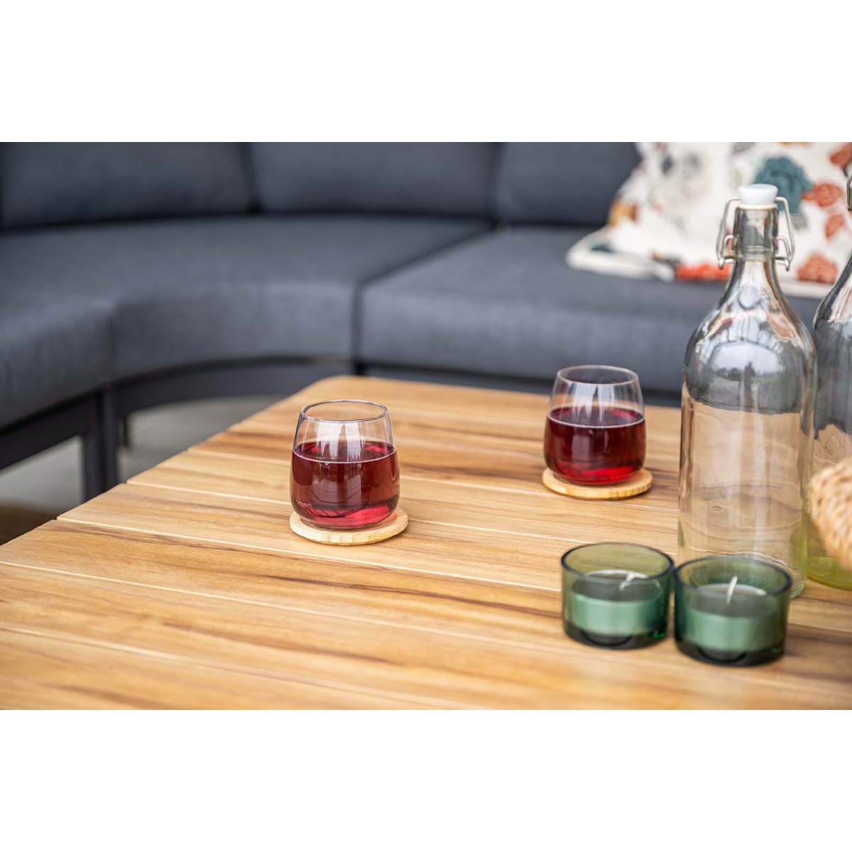 Two glasses with red liquid on coasters, two candles, and a bottle on a wooden table. A sofa is in the background.