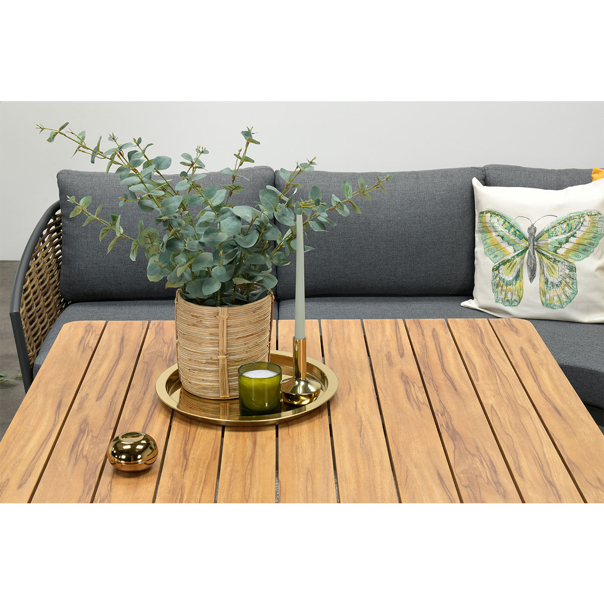 Une table en bois avec une plante, une bougie et une décoration dorée devant un canapé gris avec un coussin papillon.