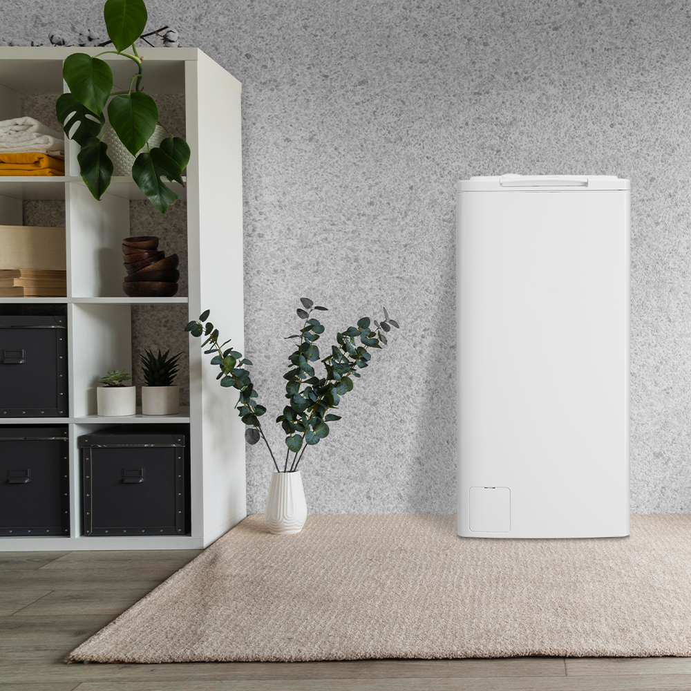 Una lavadora blanca de carga superior en una habitación con una estantería, plantas y una alfombra.