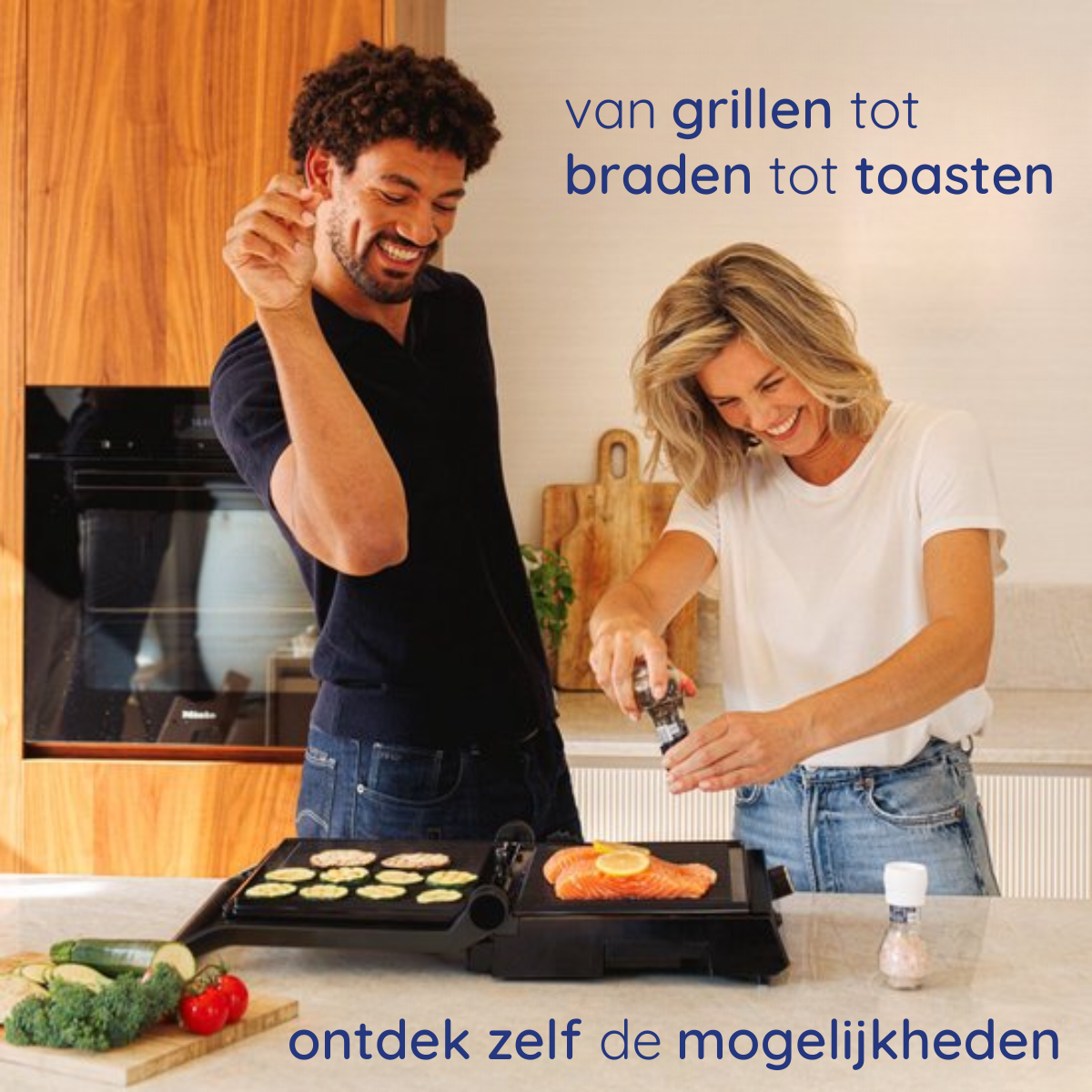 A man and woman are cooking with a grill. Various food items are on the grill. Kitchen setting is visible.