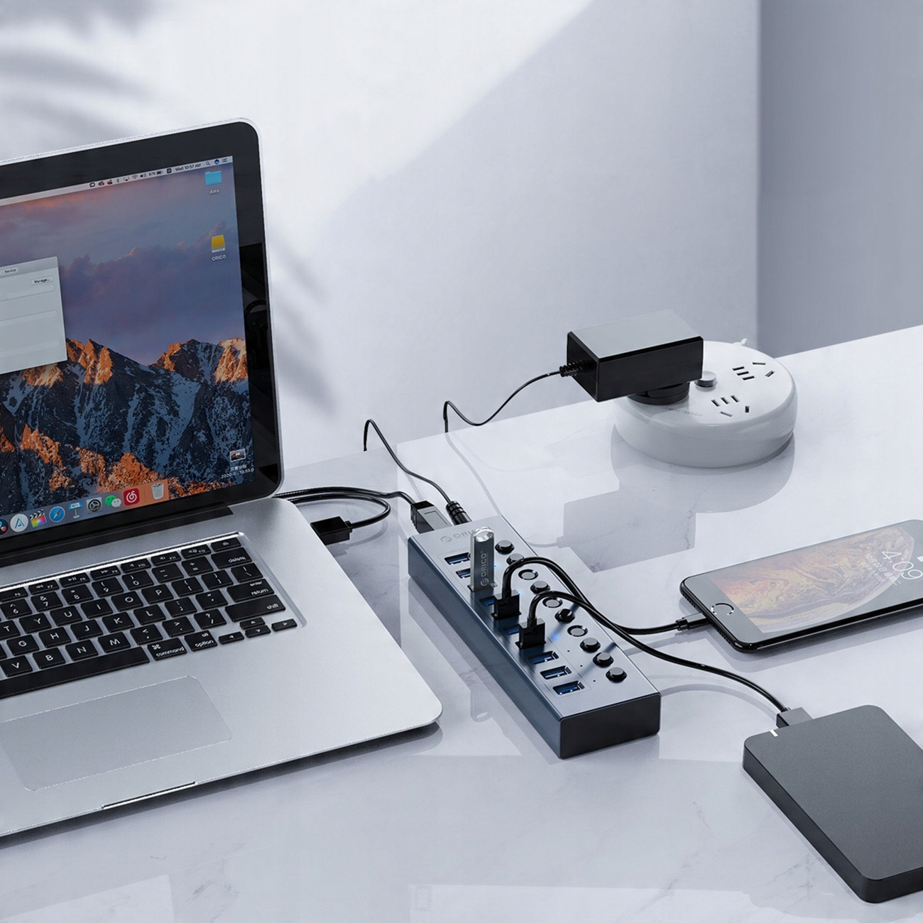 Laptop, koncentrator USB, smartfon i power bank, wszystko na białym biurku, z wieloma kablami.