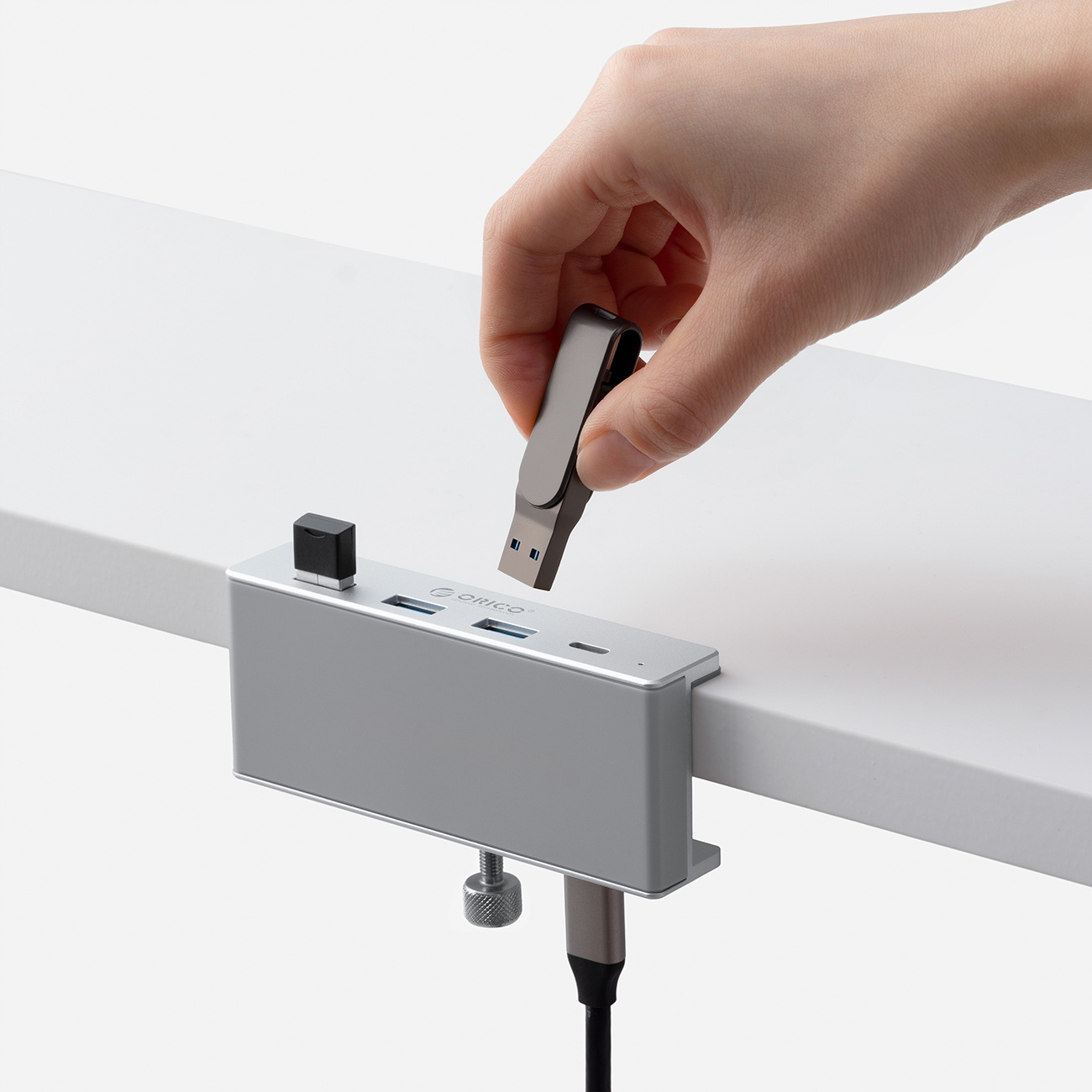 Osoba trzymająca pendrive w kierunku srebrnego huba USB przymocowanego do białego biurka.