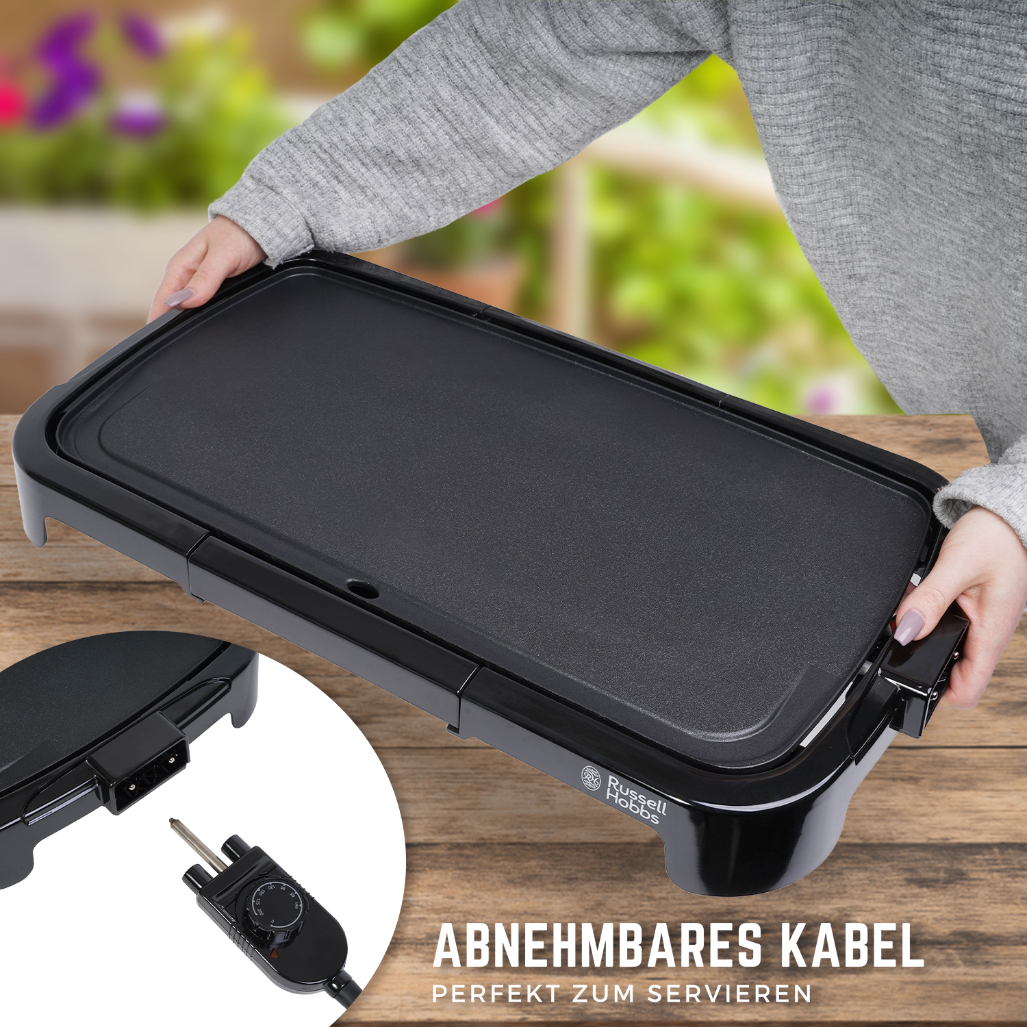 Ein schwarzer Elektrogrill mit abnehmbarem Kabel auf einer Holzoberfläche. Der Grill hat das Russell Hobbs Logo.