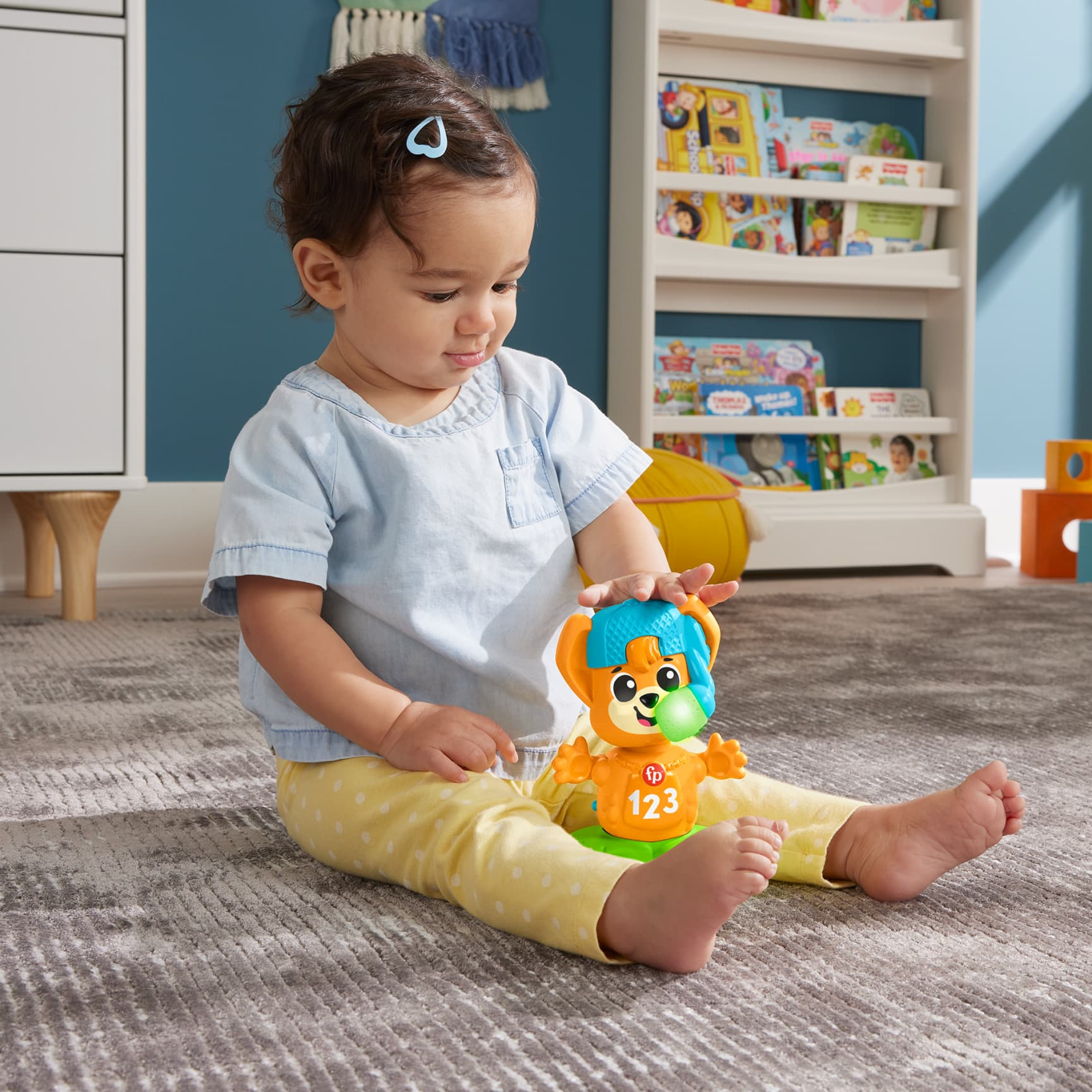 Ein Baby sitzt auf einem Teppich und spielt mit einem orangefarbenen und grünen Spielzeug mit der Aufschrift "123", im Hintergrund ein Bücherregal.