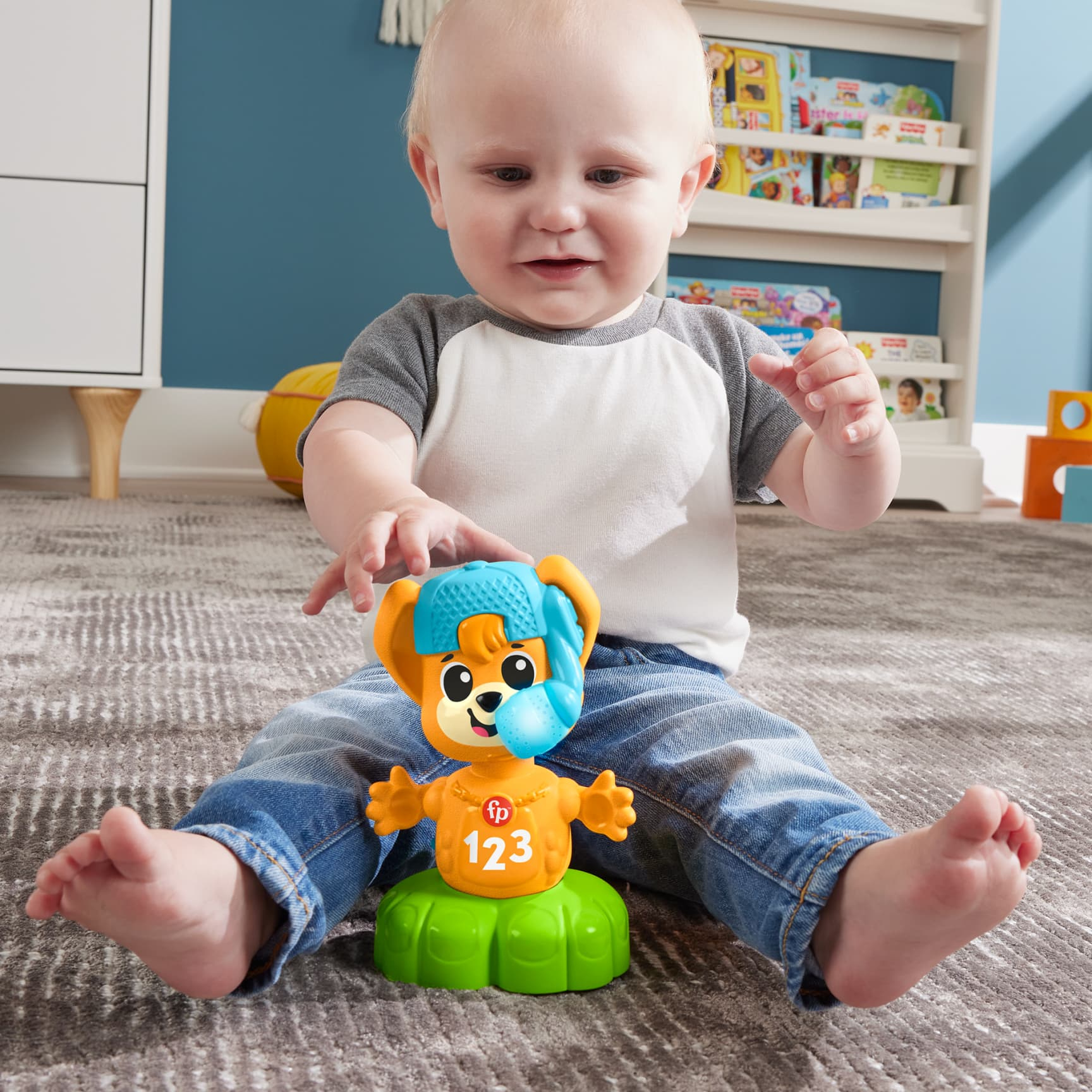 Ein Baby sitzt auf einem Teppich und spielt mit einem orangefarbenen und grünen Spielzeug mit der Aufschrift "123", im Hintergrund ein Bücherregal.