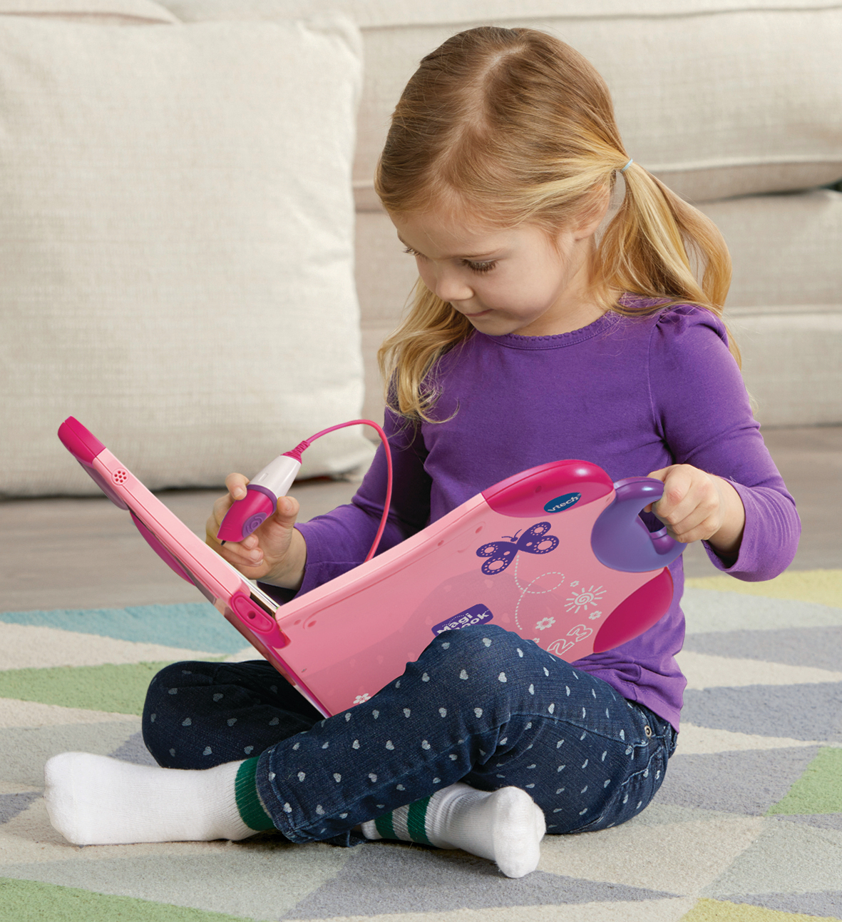 Ein Mädchen sitzt und spielt mit einem pinkfarbenen MagiBook. Sie trägt ein lila Hemd und Jeans.
