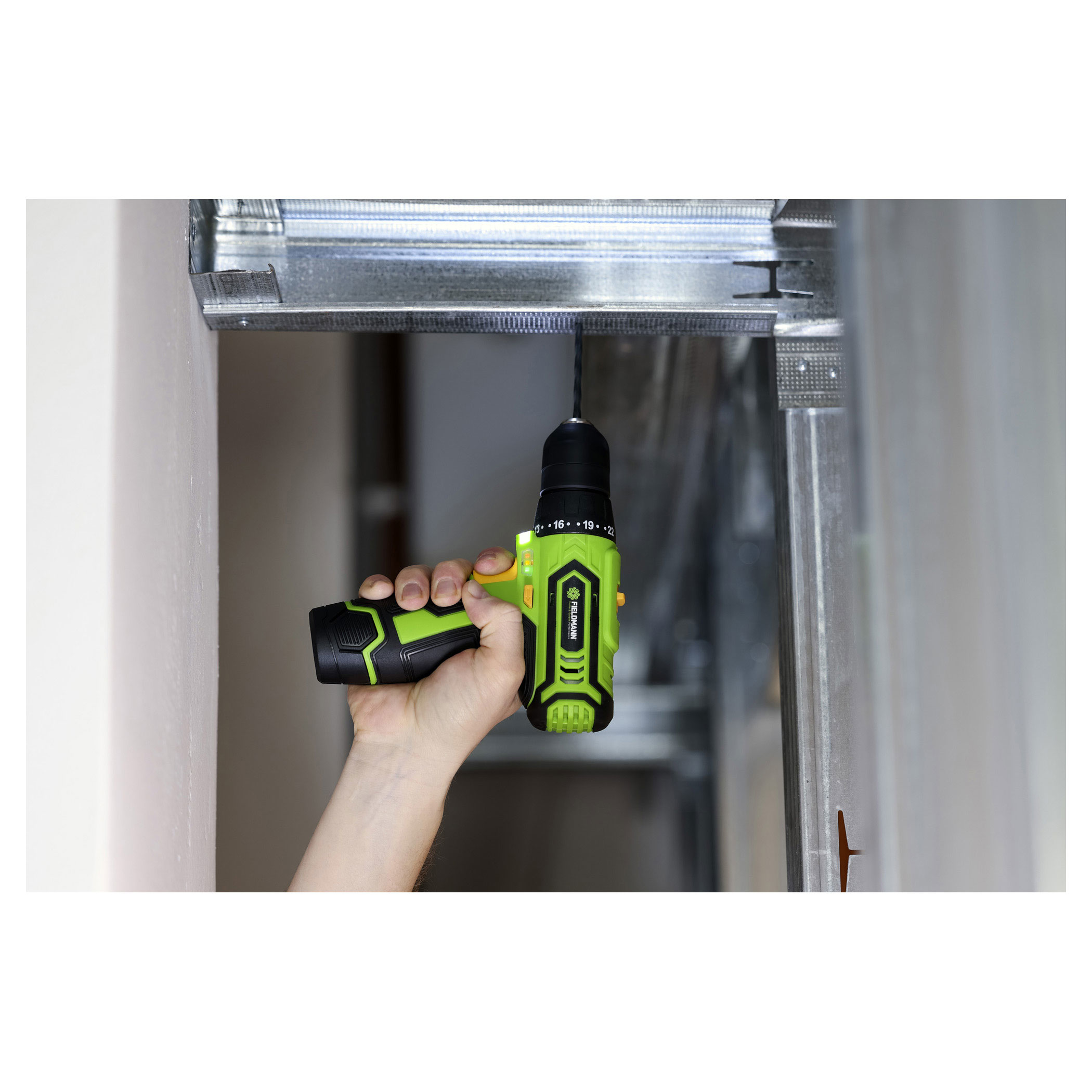 Osoba używająca zielono-czarnej wiertarki do wiercenia w metalowej ramie. Biała ściana.