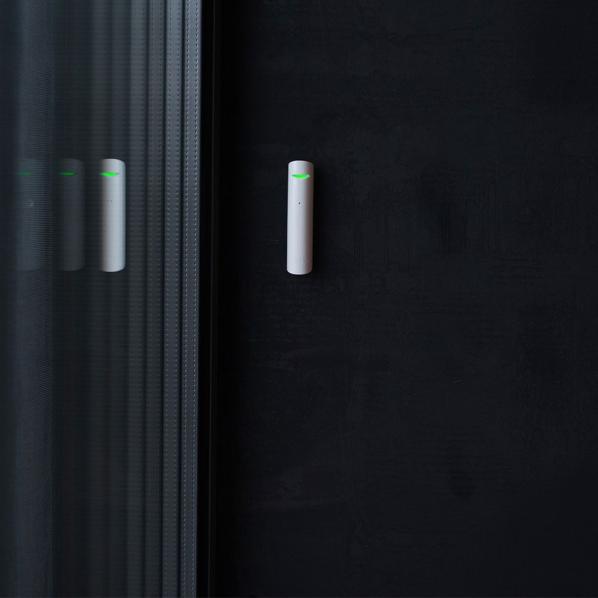 Un sensor de seguridad blanco con luz verde, montado en una pared negra con reflejo.