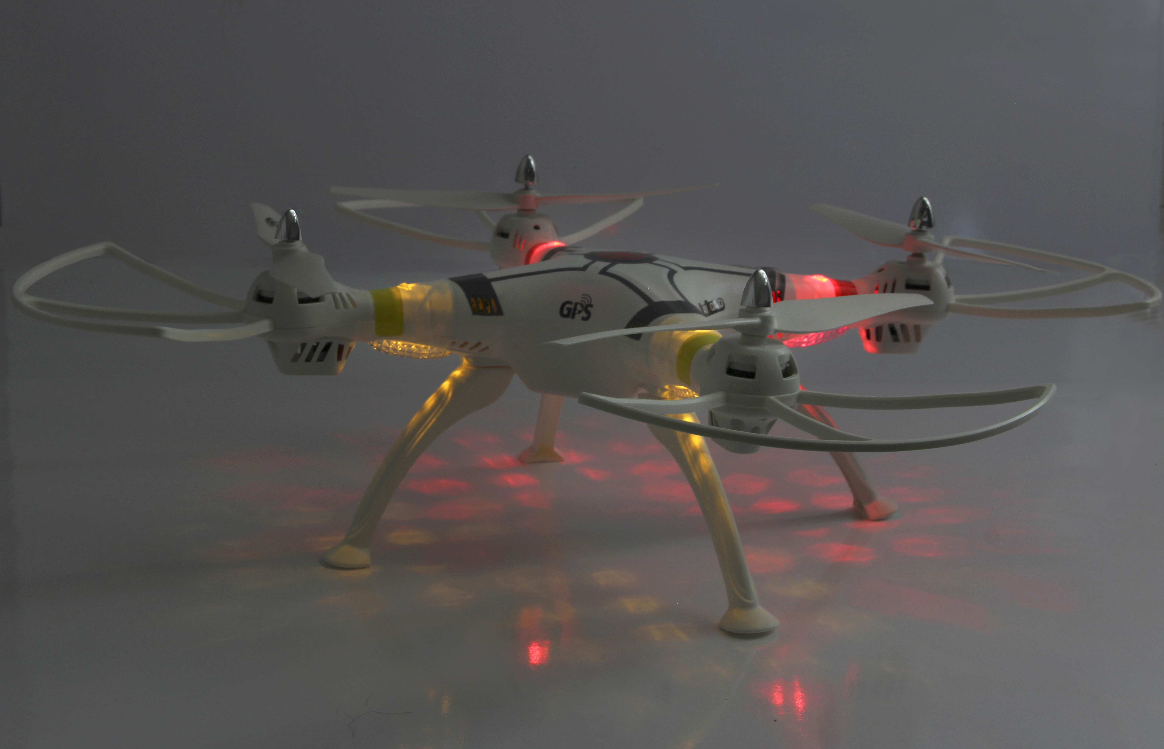 Dron z białą ramą i włączonymi światłami, oświetlającymi śmigła i korpus. Dron spoczywa na powierzchni.