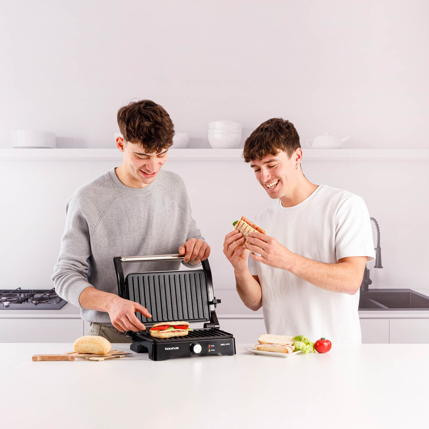 Zwei Personen, die mit einem schwarzen Grill Sandwiches zubereiten. Auf einem Tisch liegen Brot, Gemüse und fertige Sandwiches.
