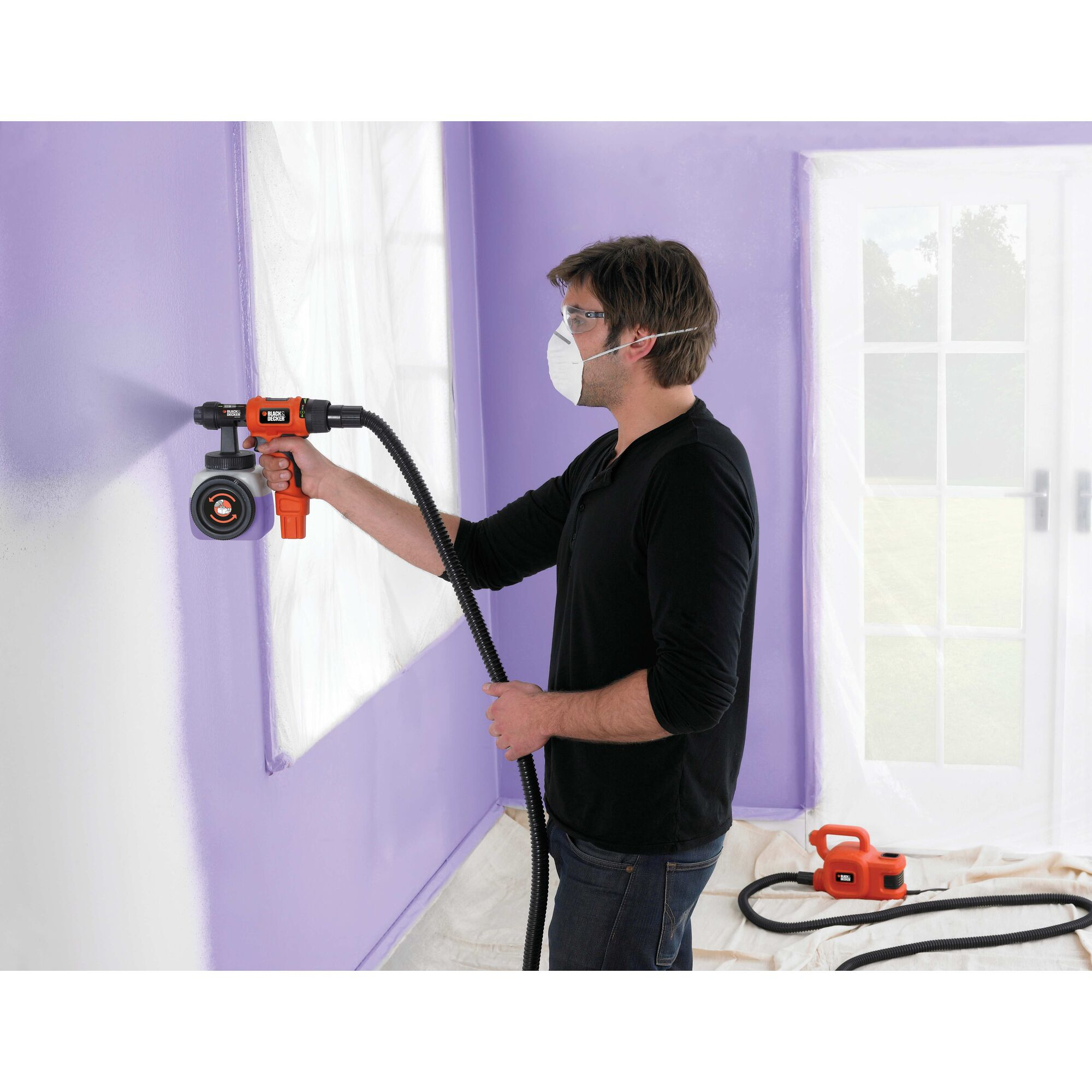 Un uomo con la mascherina spruzza un muro viola con un aerografo arancione e nero, in un ambiente interno.