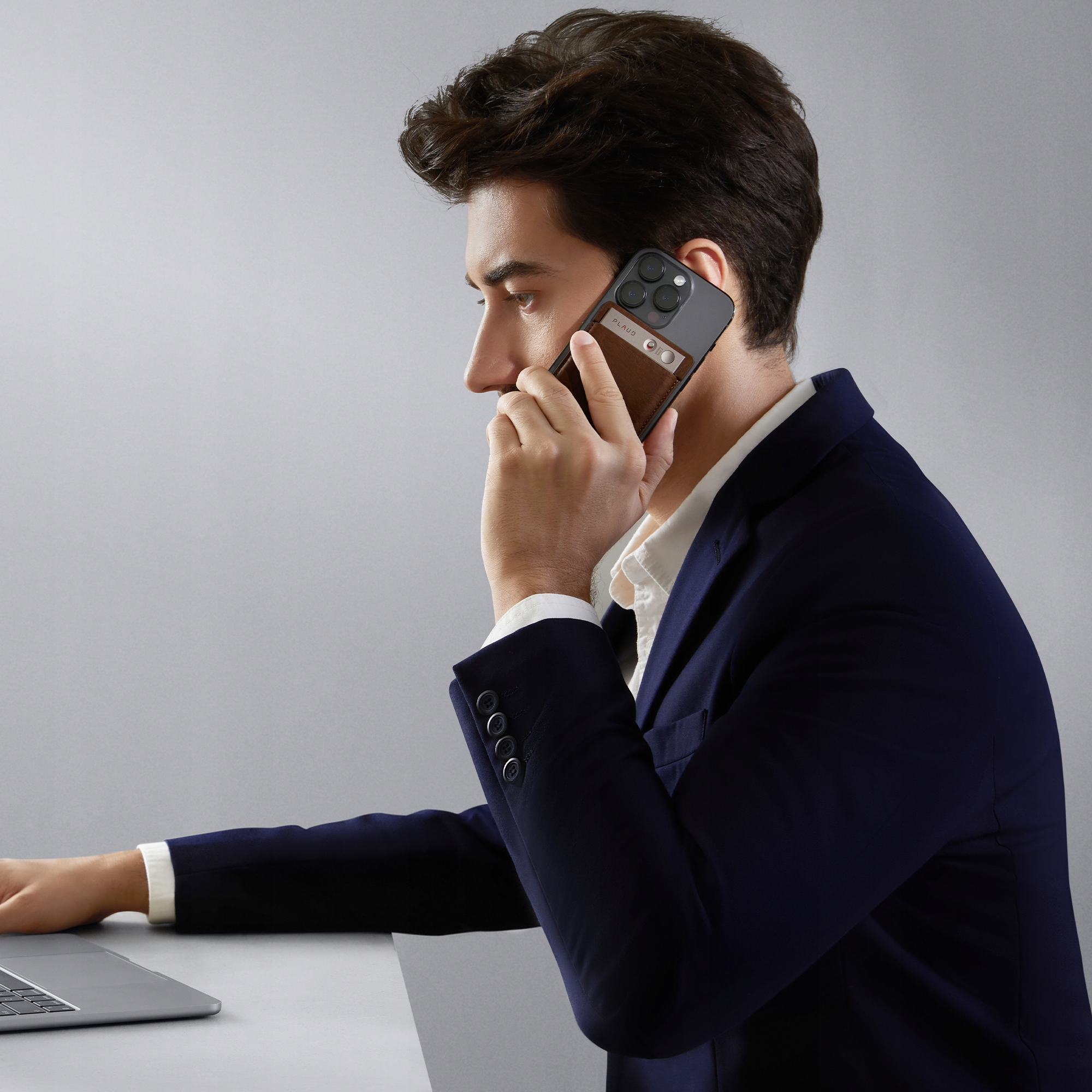 Mężczyzna w garniturze rozmawia przez telefon, pracując na laptopie. Ma telefon z etui na karty.