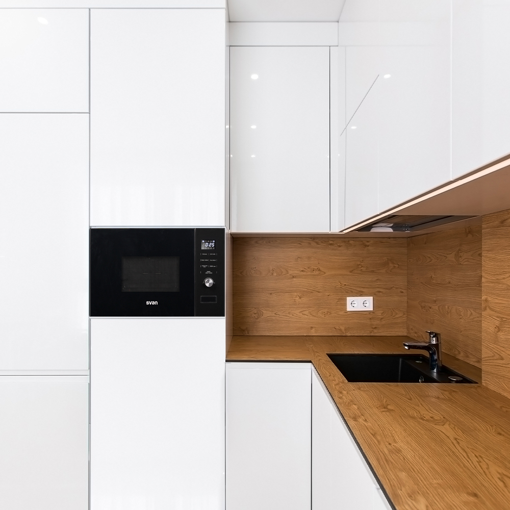 Escena de cocina moderna. Armarios blancos, encimera y salpicaderos de madera. Horno microondas y un fregadero negro. Luminoso, limpio.