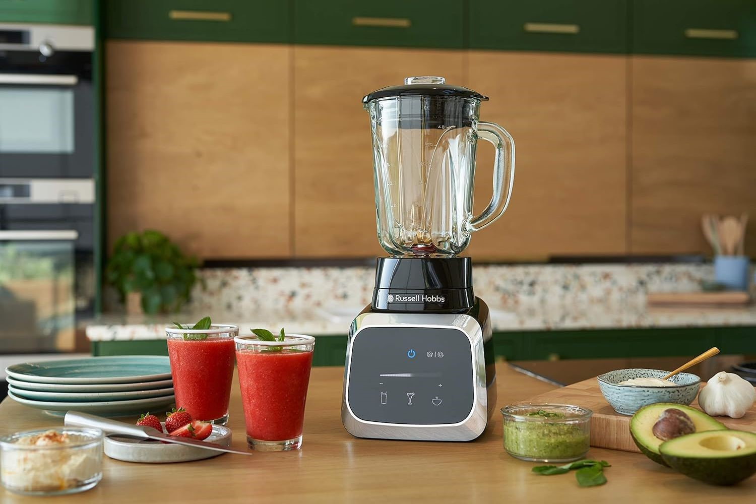Blender mit zwei Gläsern rotem Smoothie, Früchte auf der Theke. Grüne Küche im Hintergrund.