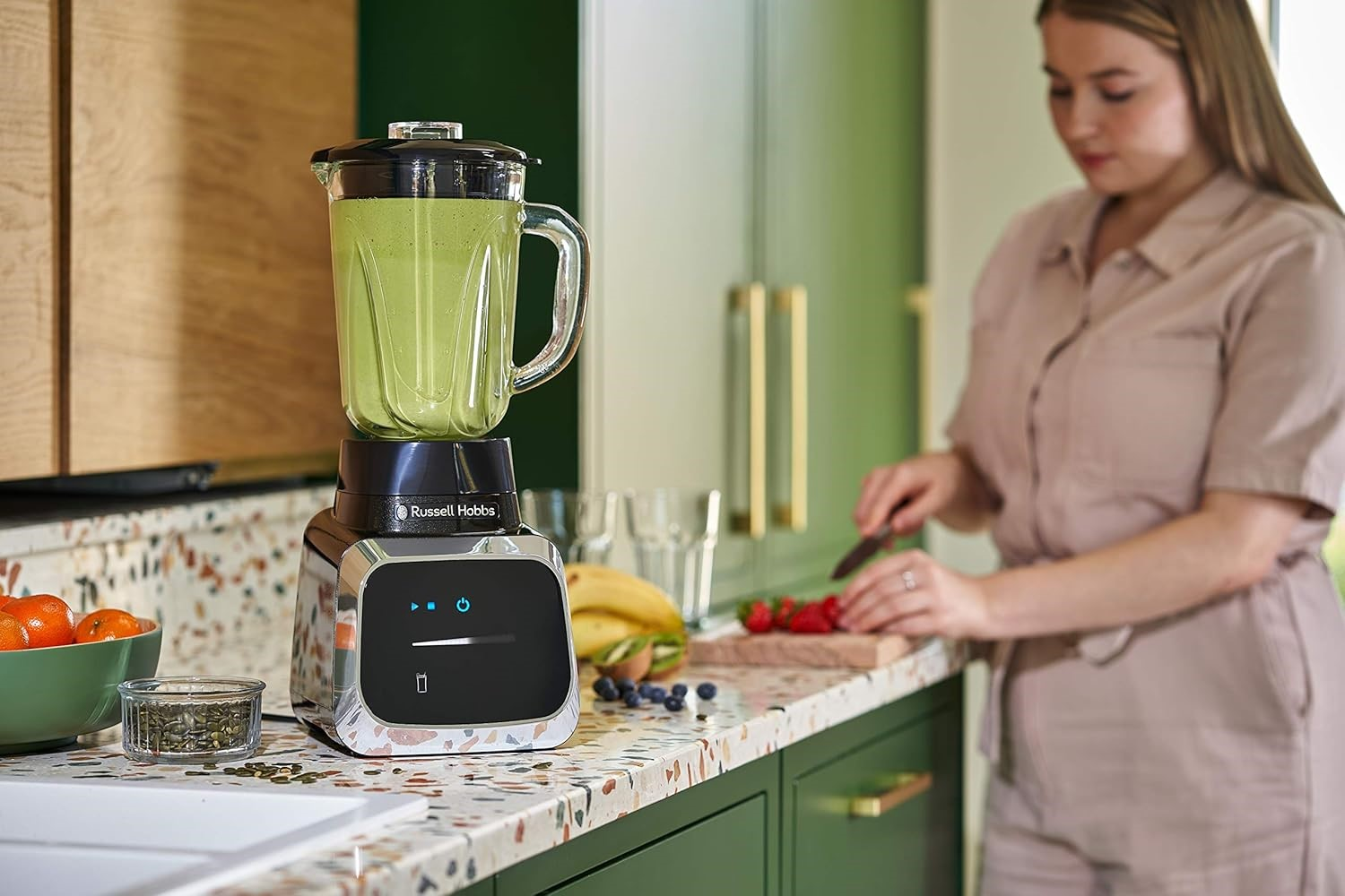 Blender mit grünem Smoothie, Frau schneidet Erdbeeren. Küche mit grünen Schränken und Holz.