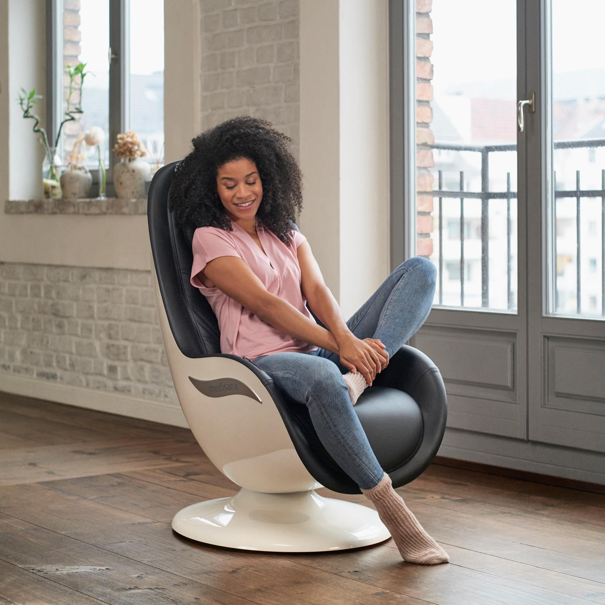 Een vrouw zit in een moderne, zwart-witte massagestoel. Ze glimlacht en ontspant zich bij een raam.