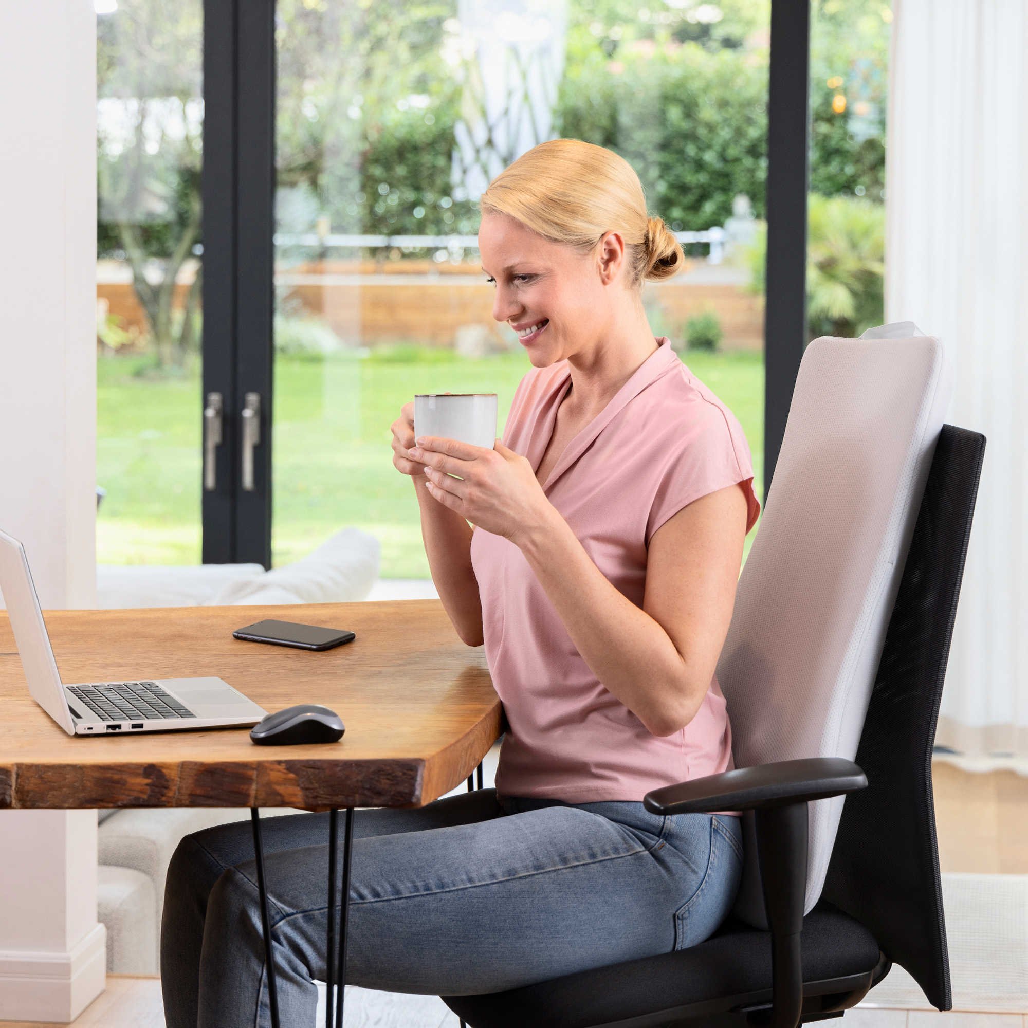 Kobieta w różowej koszuli pije z kubka przy biurku, z laptopem i podparciem pleców.