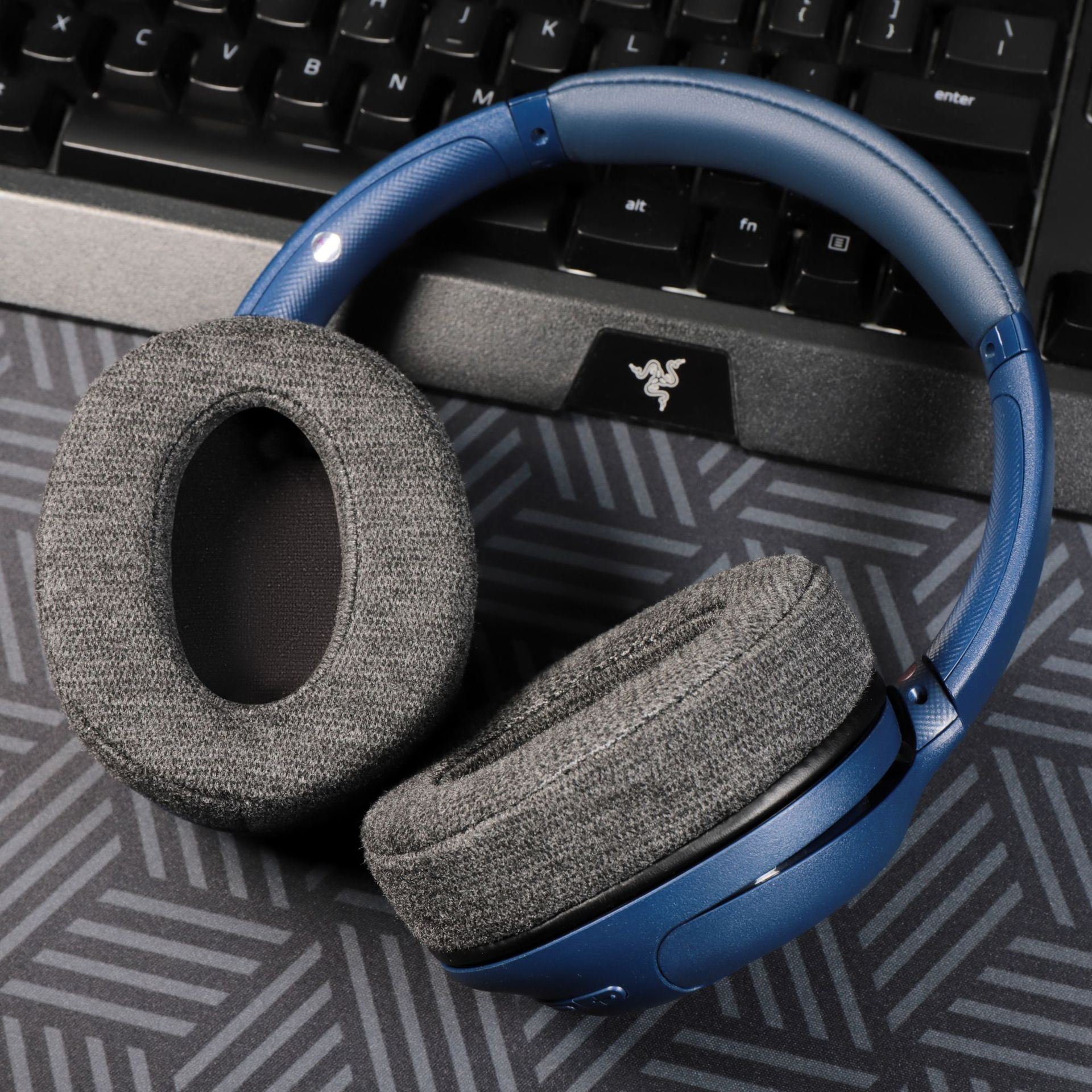 Blaue Kopfhörer auf einer gemusterten Oberfläche. Im Hintergrund befindet sich eine Tastatur. Razer-Logo sichtbar.