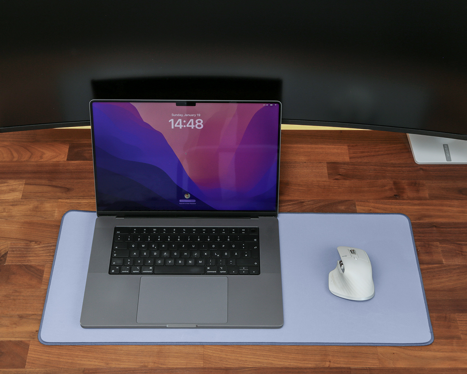 Ein Laptop, eine Maus und ein Mousepad auf einem Holztisch. Im Hintergrund ein Display.