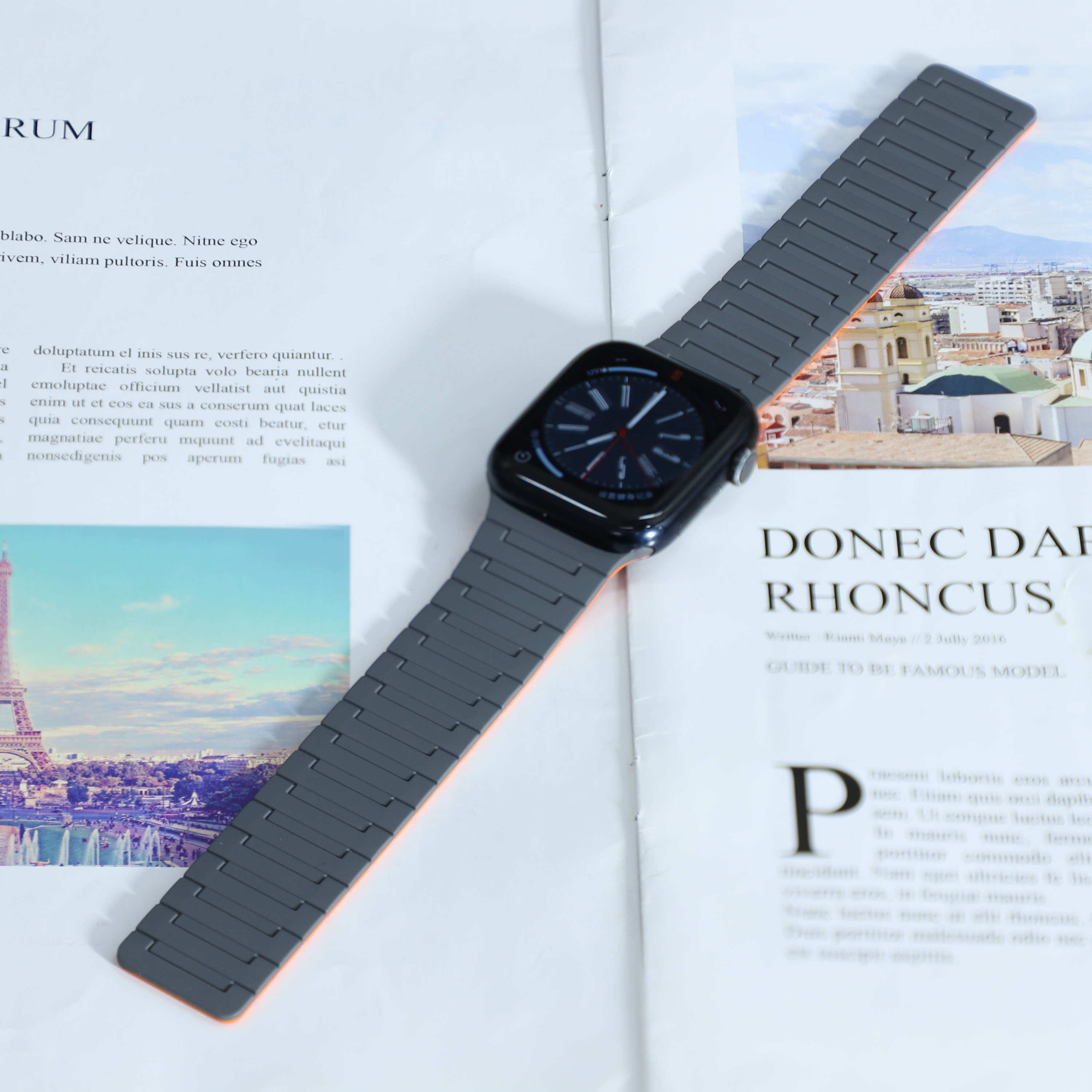 Smartwatch mit grauem Armband und schwarzem Zifferblatt, auf einer bedruckten Zeitschrift liegend. Hintergrund mit Gebäuden.