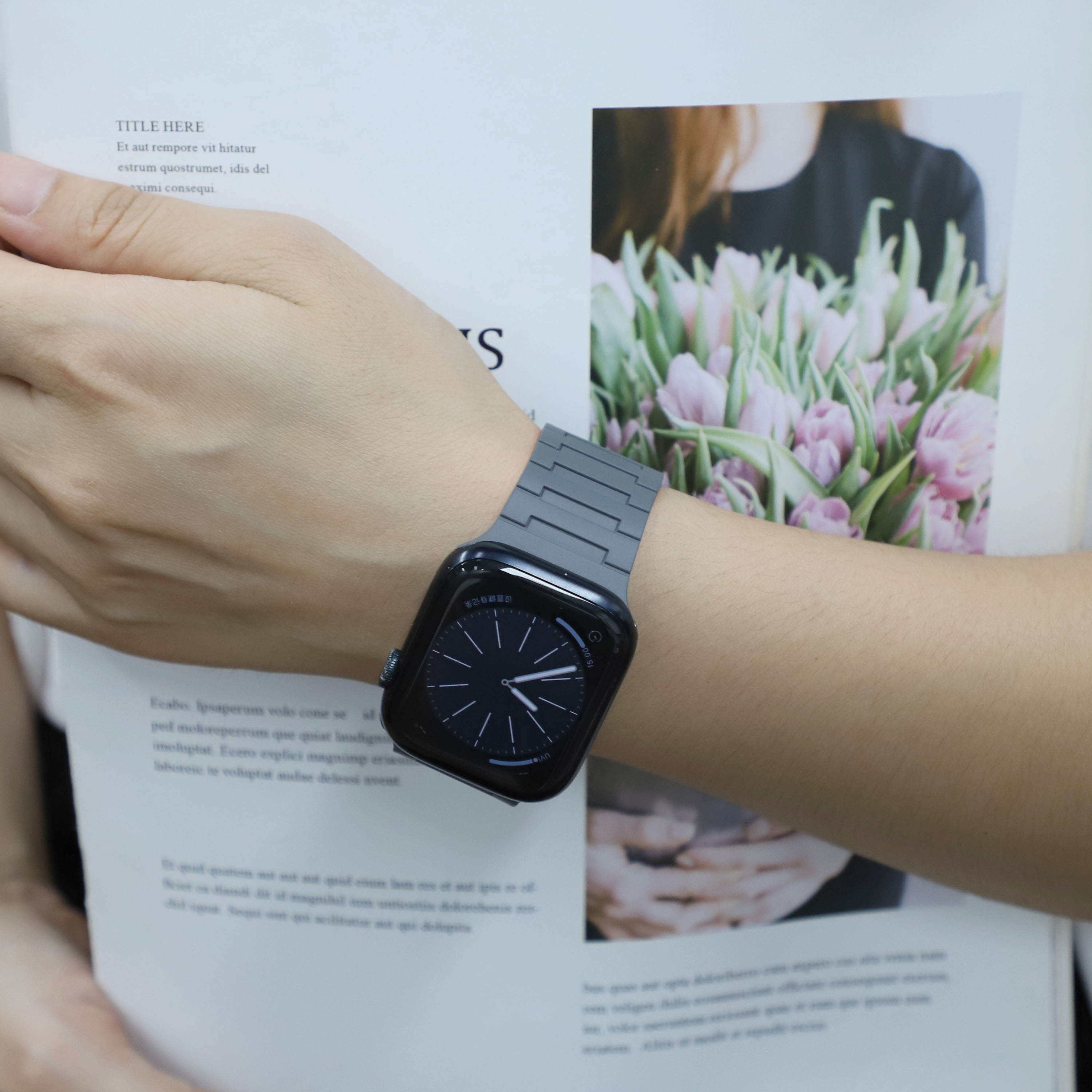 Person mit schwarzer Apple Watch und grauem Armband. Offenes Buch mit Text im Vordergrund, Blumen im Hintergrund.