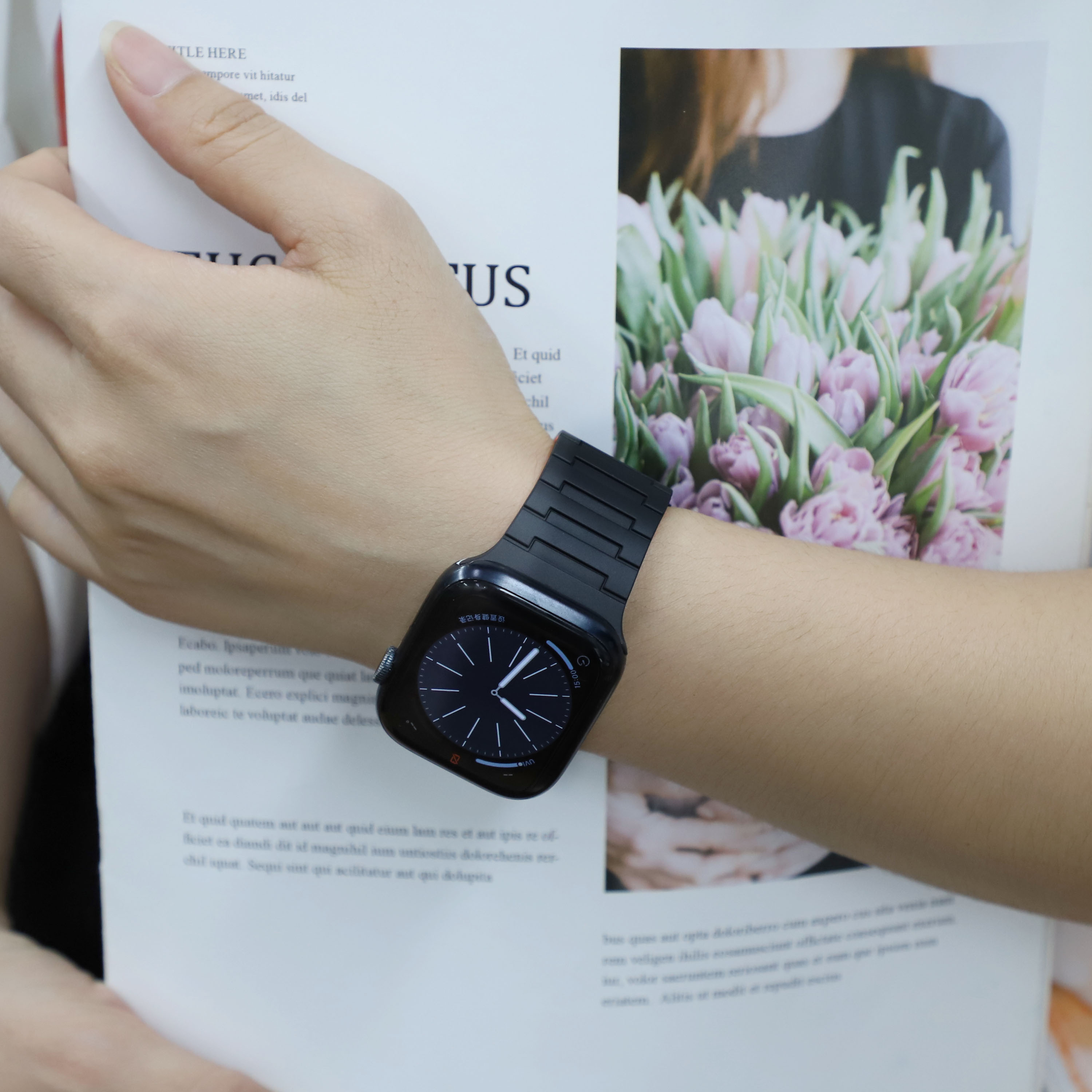 Person mit schwarzer Apple Watch und schwarzem Armband. Offenes Buch mit Text und Blumen im Hintergrund.