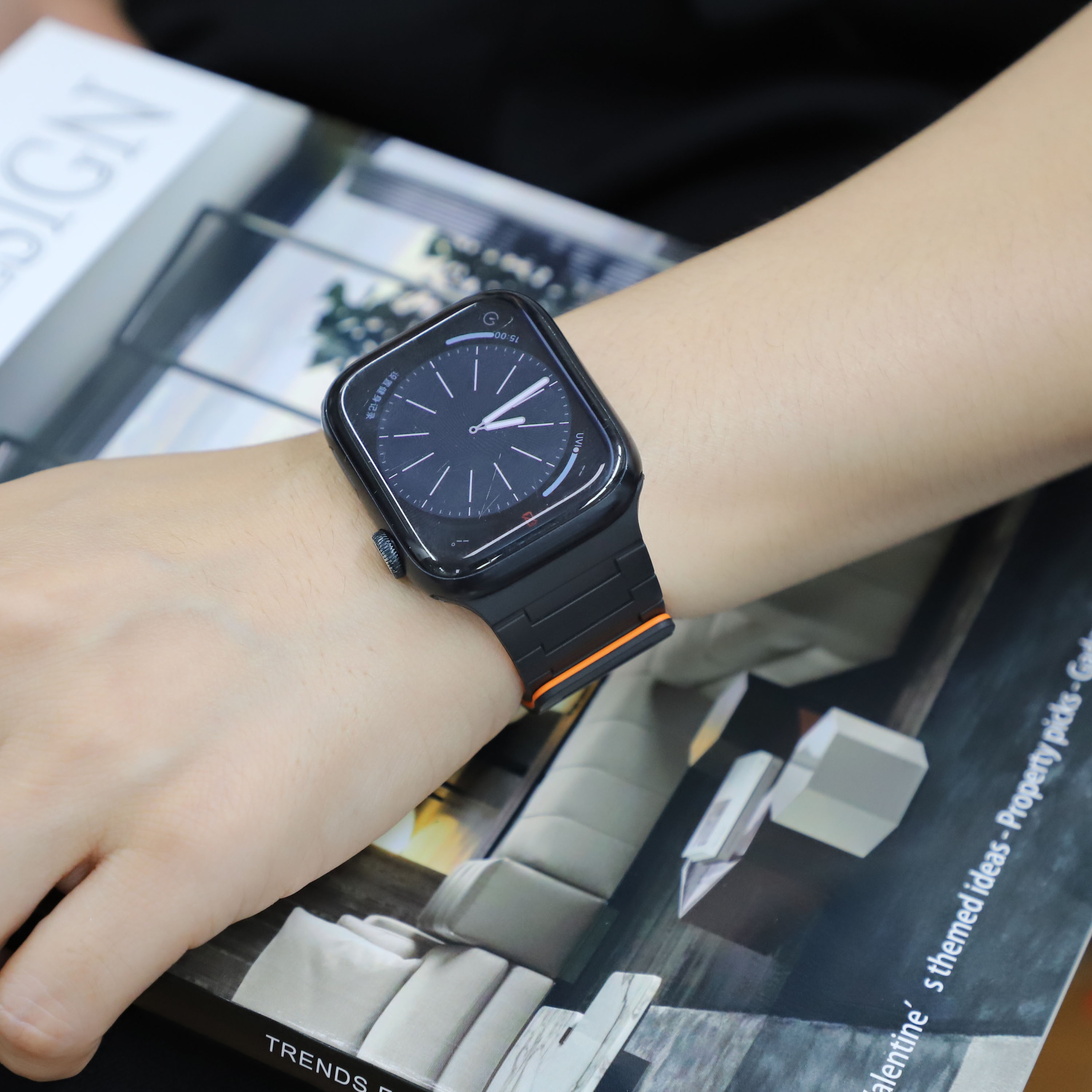 Person trägt schwarze Smartwatch mit schwarz-orangem Armband. Zifferblatt sichtbar. Hintergrund unscharf.