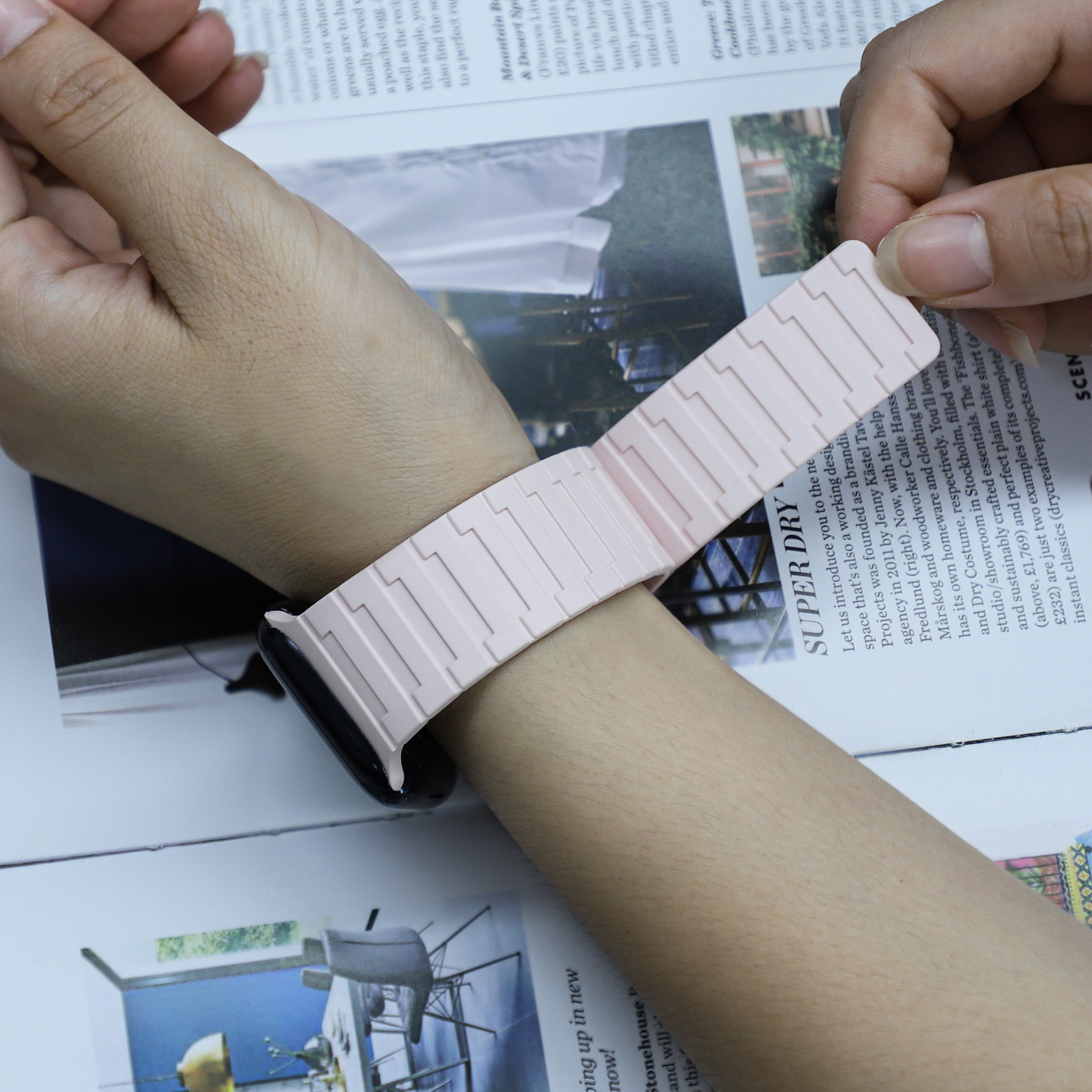 Ein Handgelenk mit einem pinkfarbenen Uhrenarmband. Die Uhr ist nicht sichtbar, im Hintergrund ein Magazin.