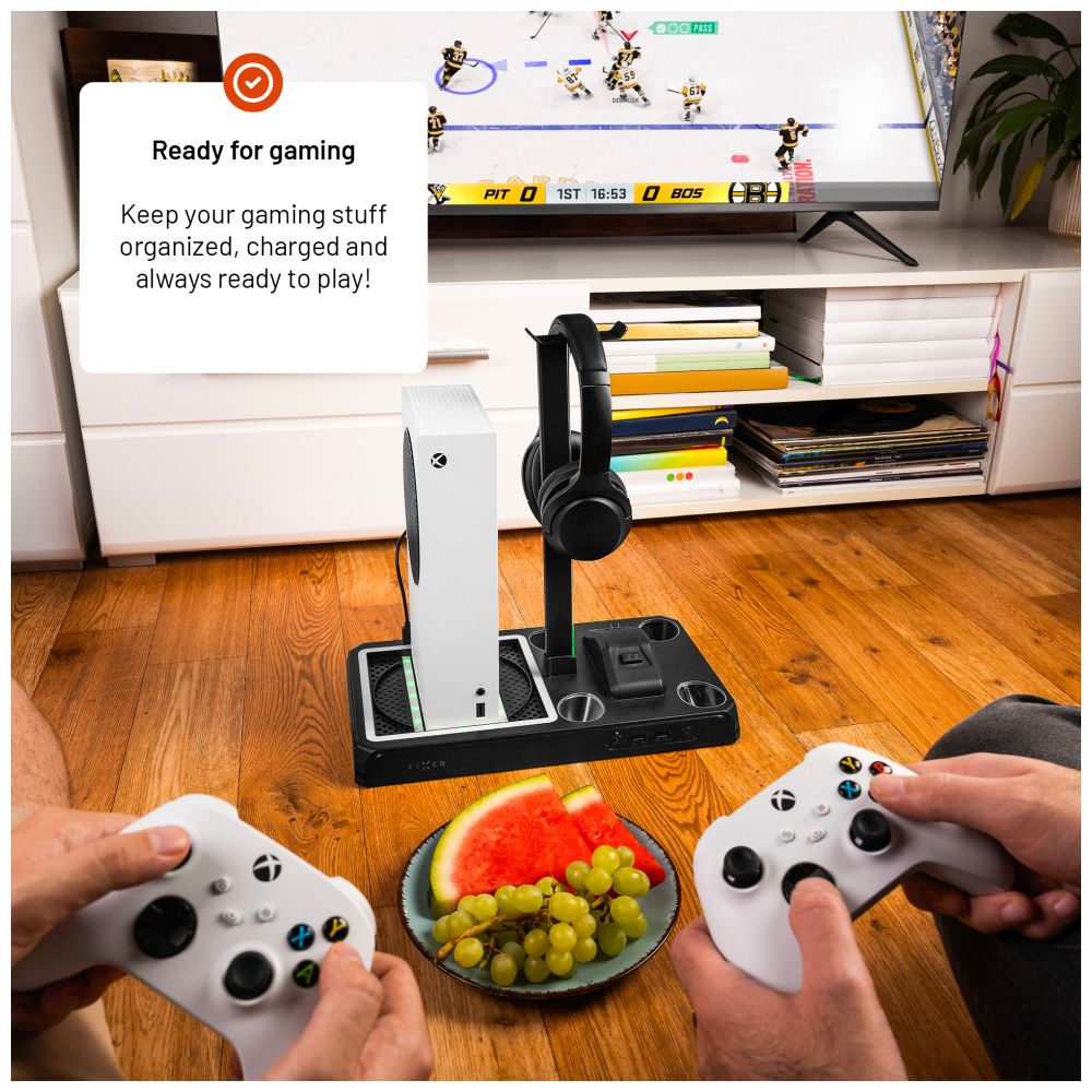 Hände halten Controller, Tisch mit Konsole und Essen; TV im Hintergrund.