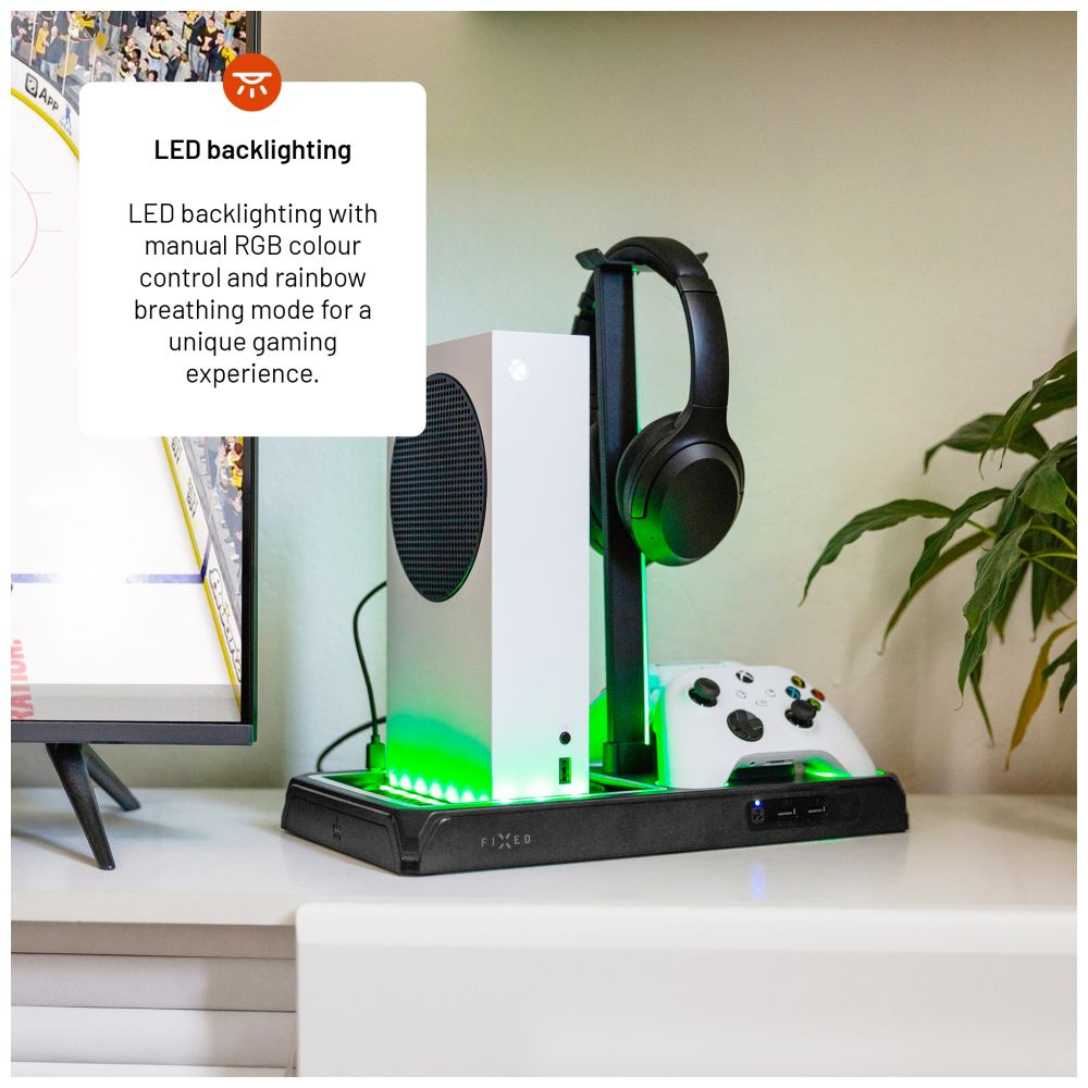 Weiße Spielkonsole mit grünen LED-Leuchten, Kopfhörern und Controller auf einem schwarzen Ständer. Pflanze und Fernseher im Hintergrund.